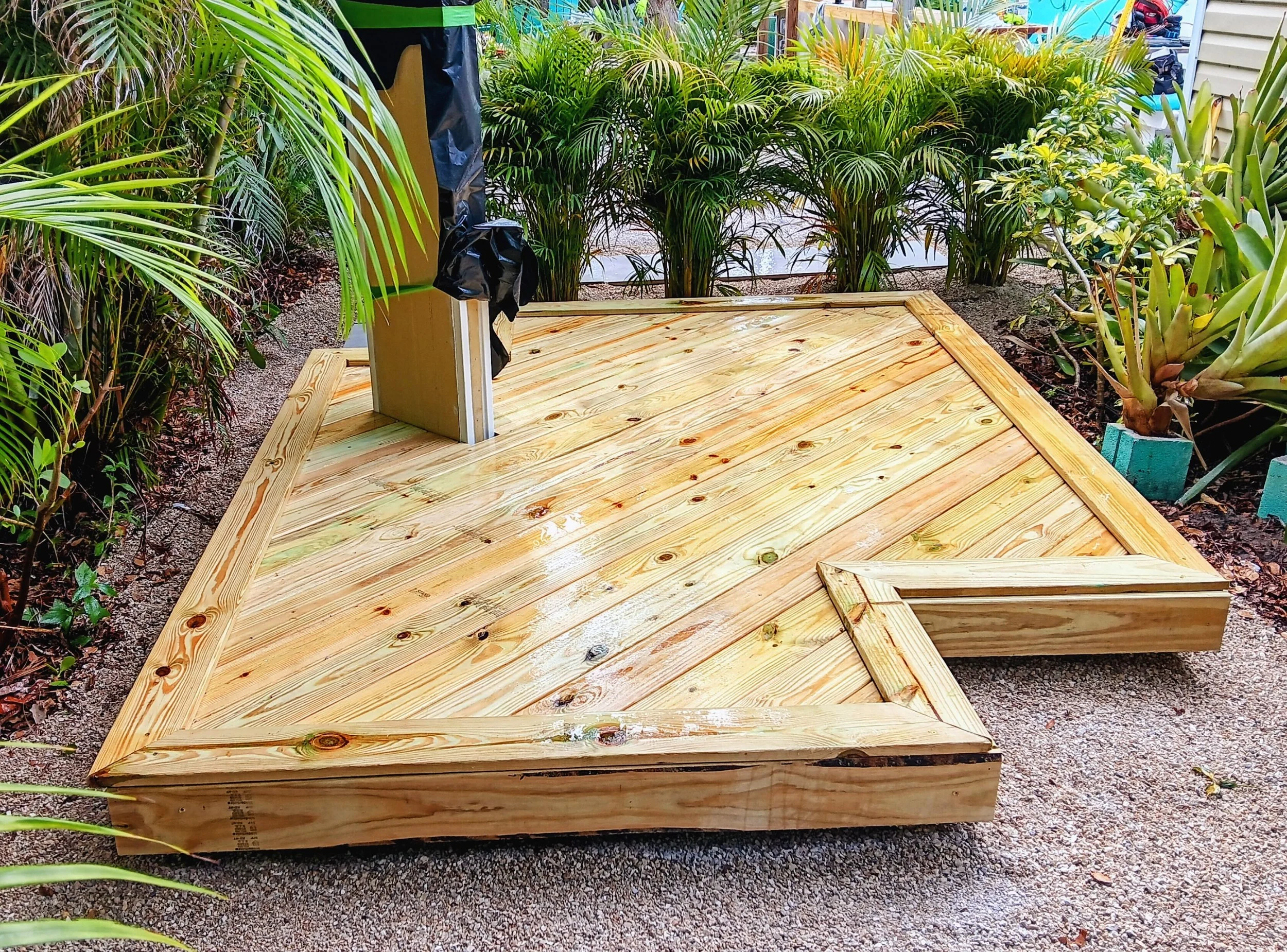 A wooden deck under construction with a corner and a step, surrounded by lush green tropical plants, with a house or building in the background.