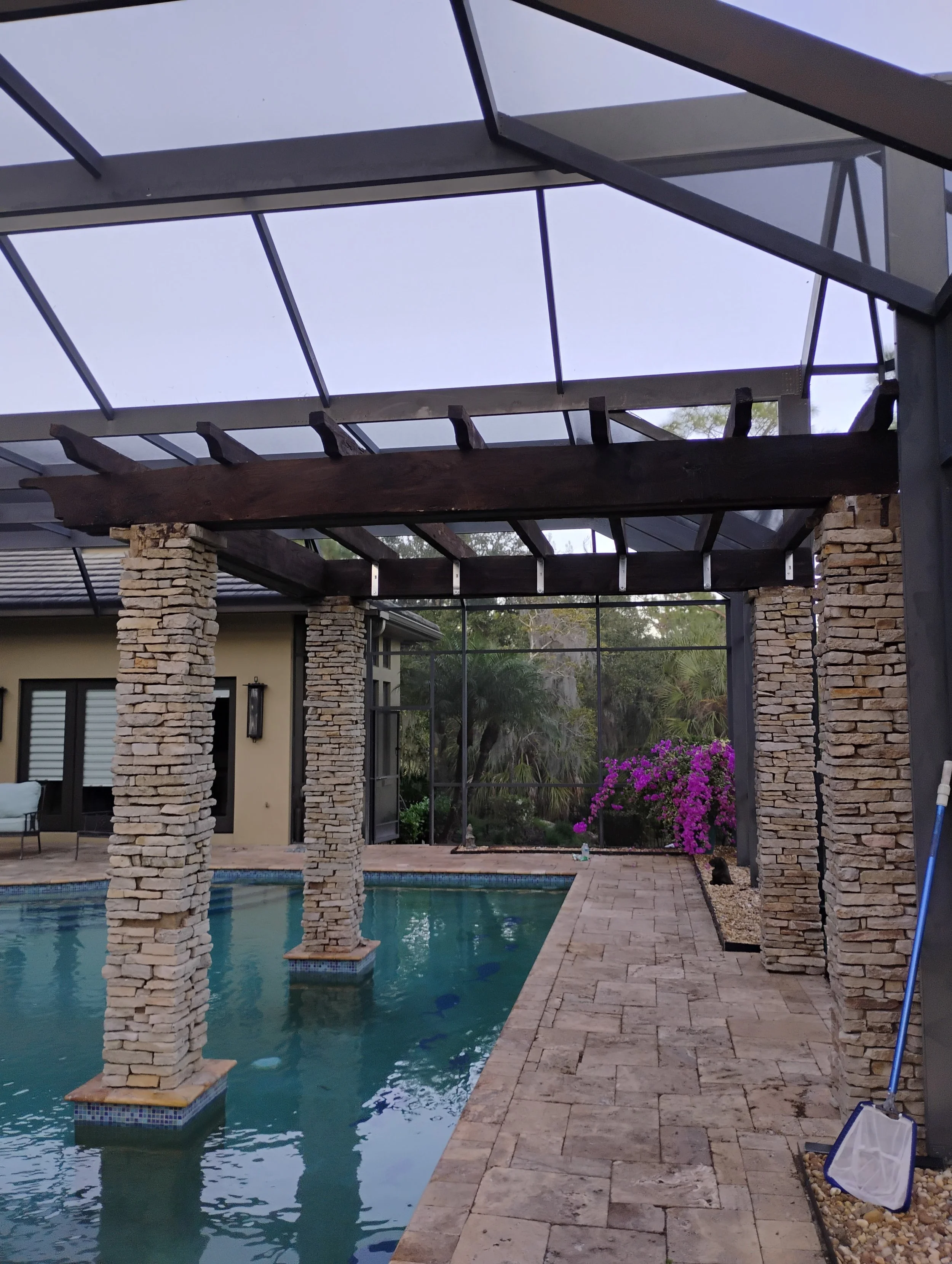 A backyard pool area with stone pillars supporting a wooden pergola, with a screened enclosure overhead. There are flowering bushes, a patio with a towel on a chair, and cleaning tools near the edge of the pool.