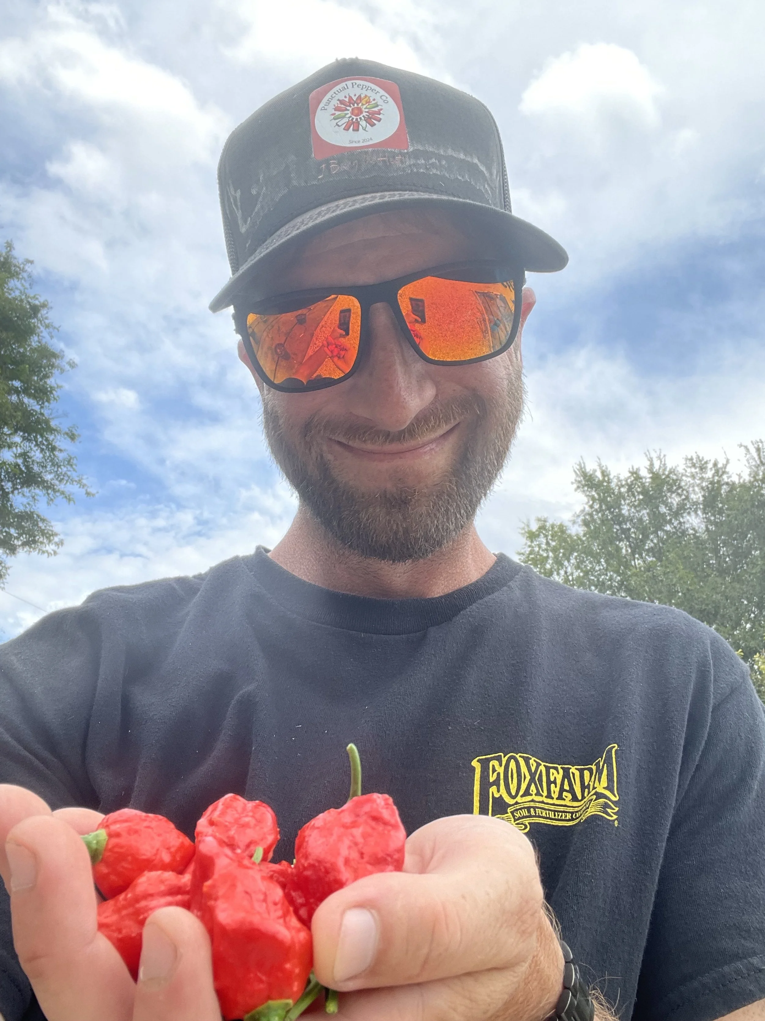 A man wearing sunglasses, a black cap with a patch that reads 'Duchual Pepper Co', and a black t-shirt with 'FOX FARM' printed on it, is smiling and holding a handful of red chili peppers outdoors with a partly cloudy sky and trees in the background.