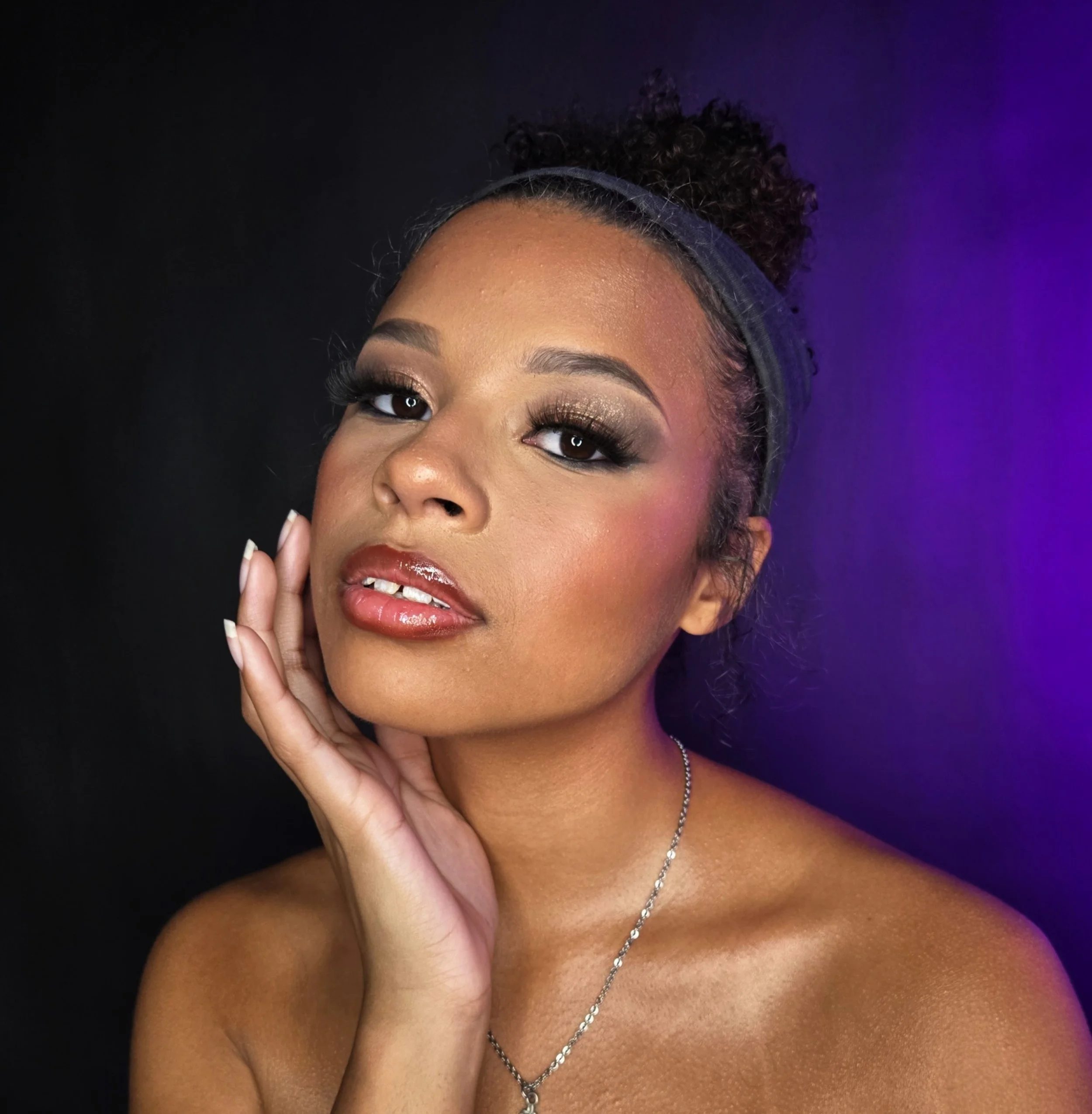 A young woman with curly hair tied back with a headband, wearing makeup with bold eye makeup, glossy lips, and a silver necklace, poses with her hand lightly touching her face against a dark background with purple lighting effects.