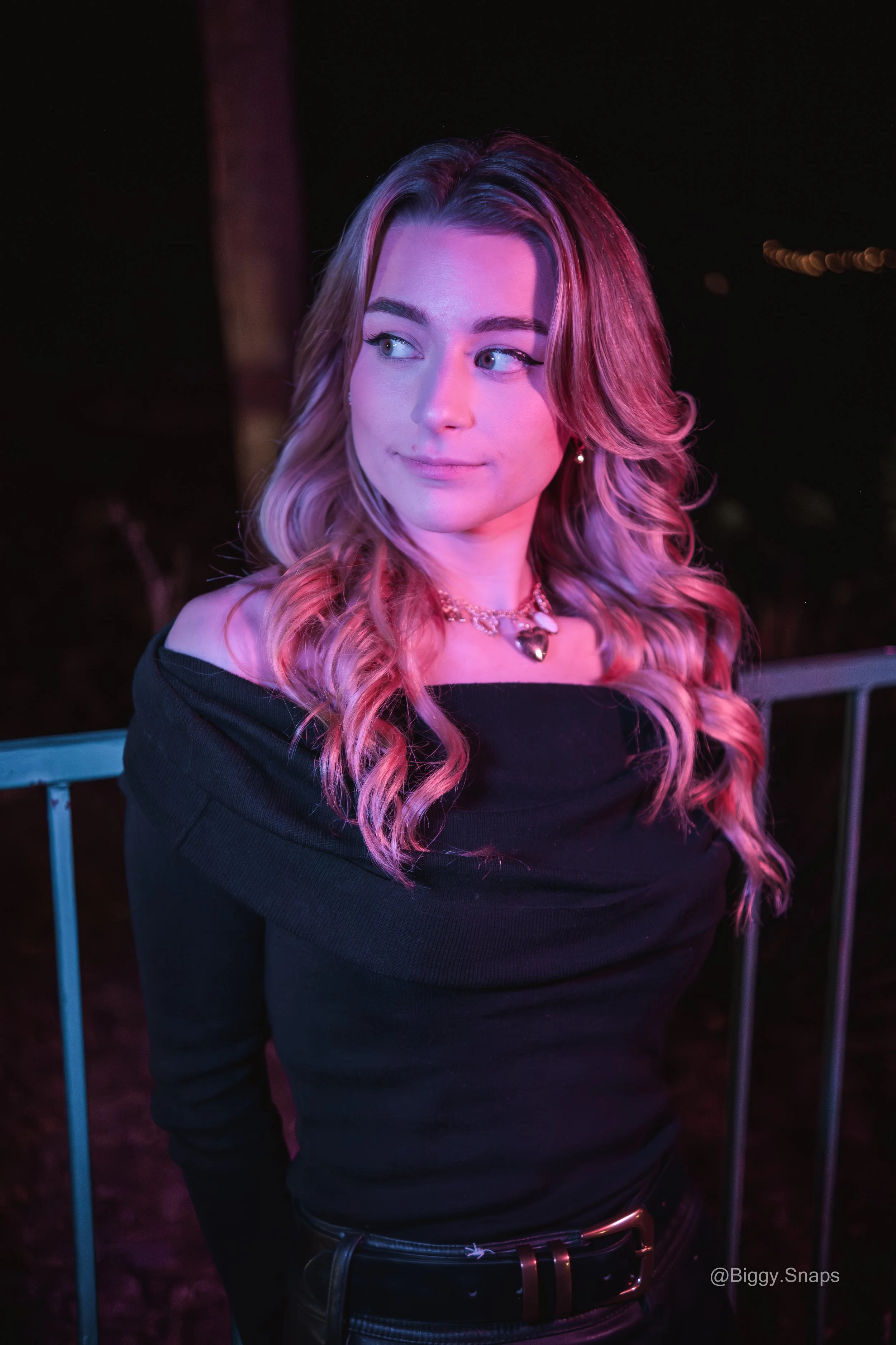 A young woman with wavy blonde hair poses at night, illuminated by pink and purple lighting, wearing a black off-the-shoulder top and a silver heart-shaped necklace.