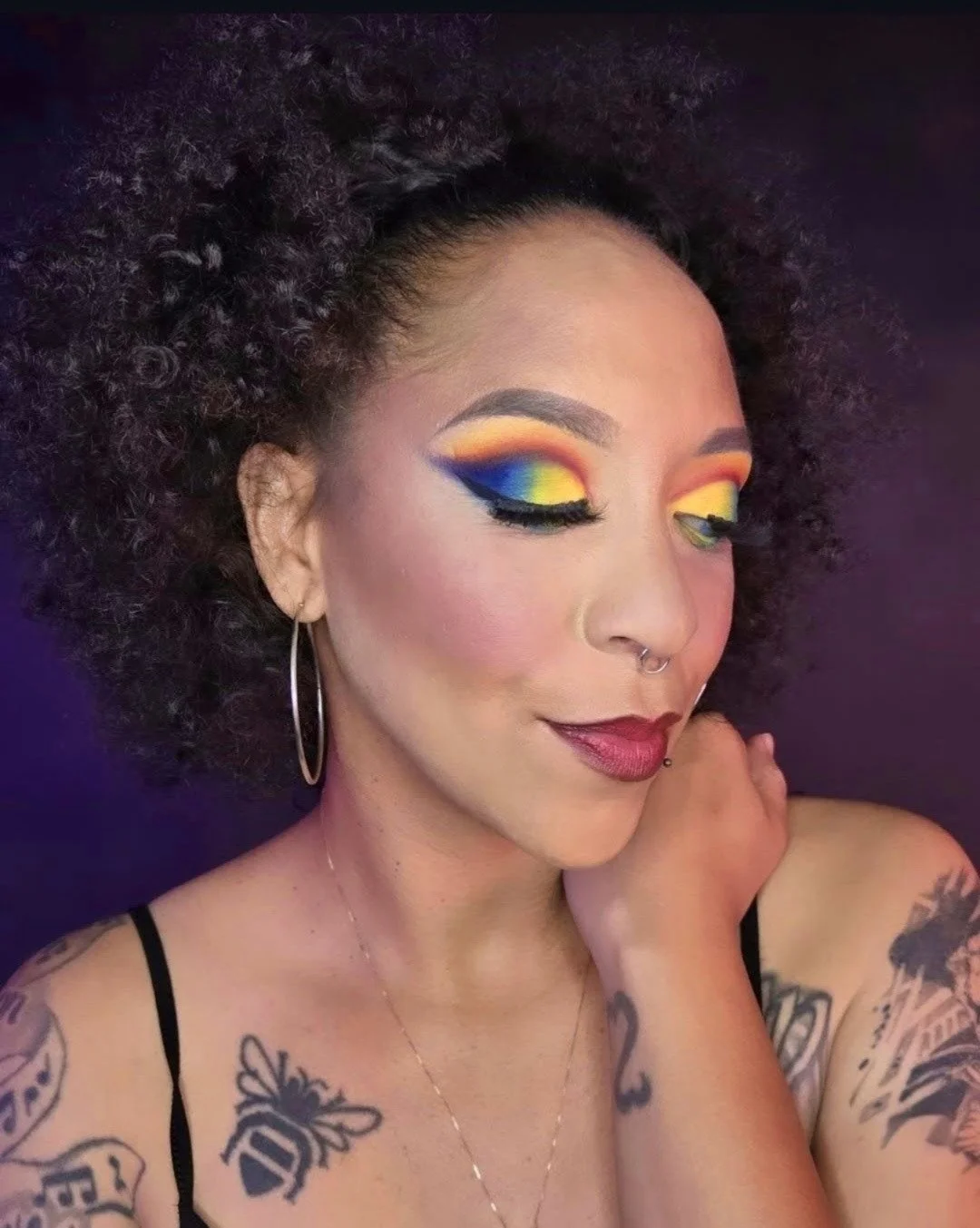 A woman with colorful rainbow eyeshadow, black eyeliner, and dark red lipstick. She has tattoos on her shoulders and wears large hoop earrings, a ring on her nose, and a small facial piercing. Her curly hair is voluminous.