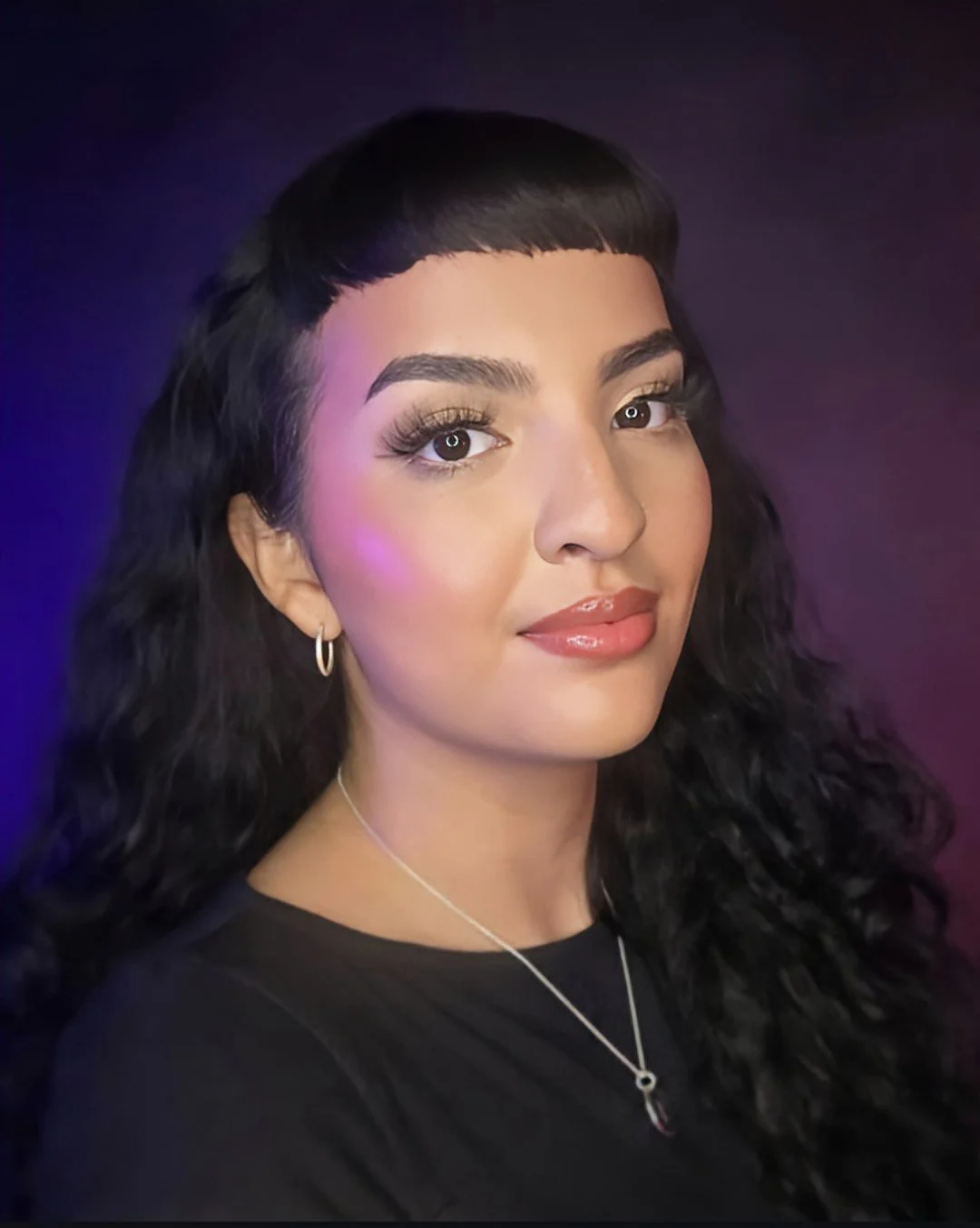Portrait of a young woman with dark curly hair, wearing makeup, hoop earrings, and a black top, against a dark background with purple and blue lighting.