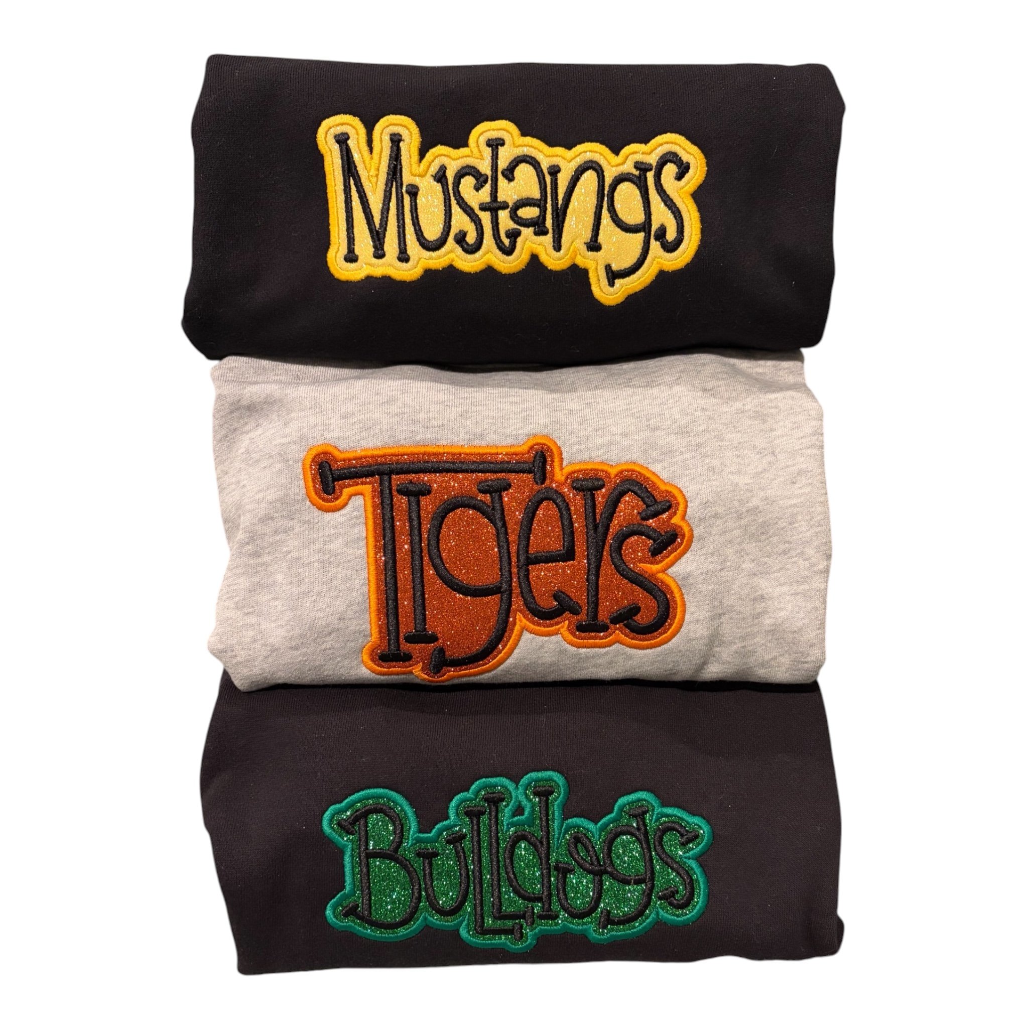 Three folded black, gray, and black sweatshirts with colorful embroidered text reading 'Mustangs,' 'Tigers,' and 'Dolphins'.