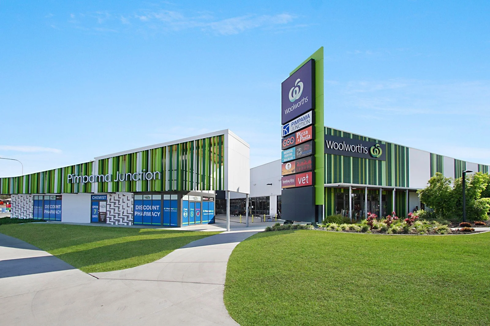 Exterior view of Pimpama Junction shopping center with green and white striped building and large signage, including Woolworths, pharmacy, and other stores, on a sunny day with a well-maintained grassy area in the foreground.
