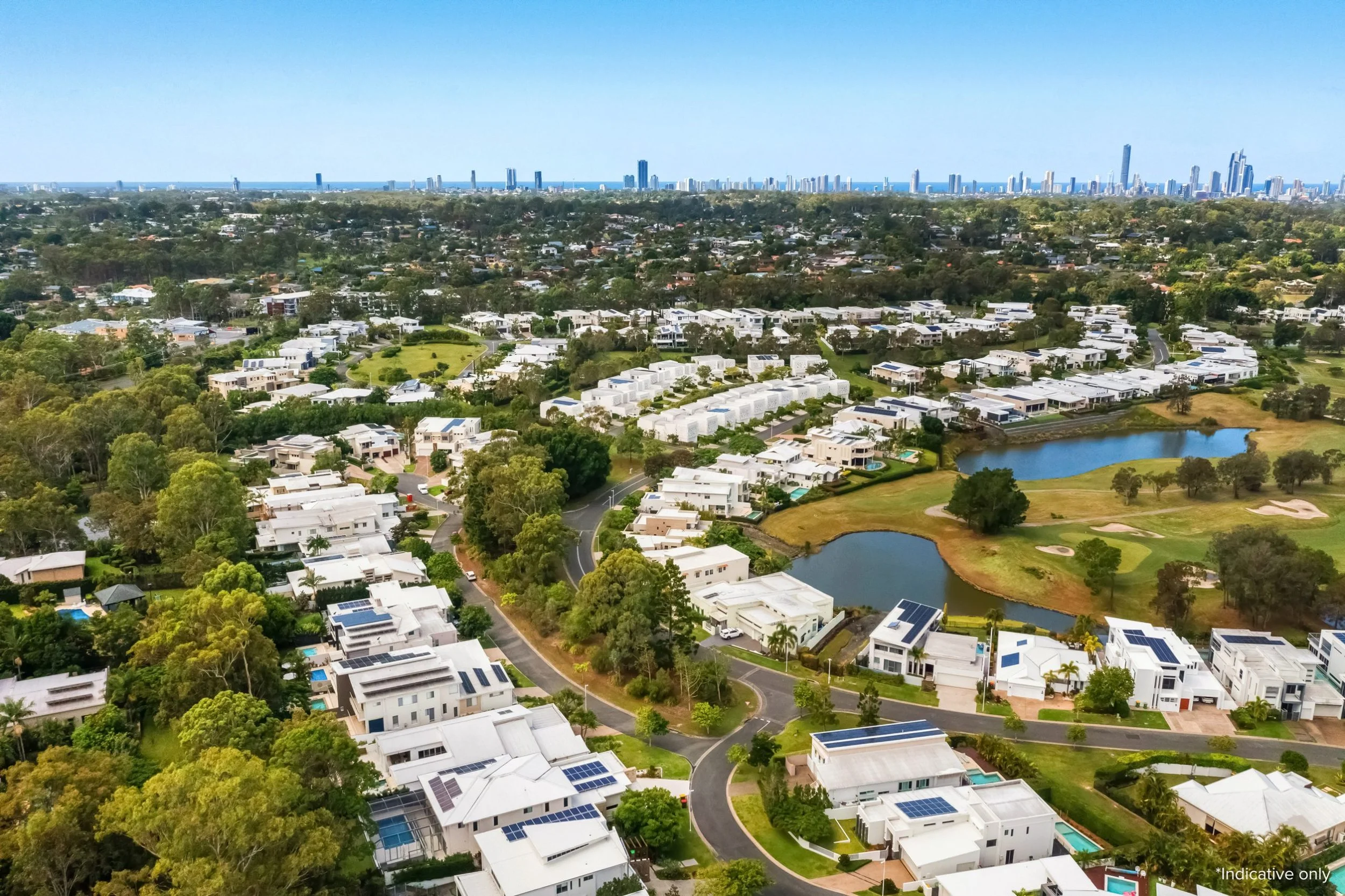 Aerial view of a suburban neighborhood with modern white houses, green trees, winding roads, a pond, and a city skyline in the distance.