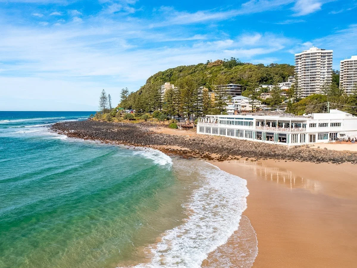 Beach with turquoise water, sandy shore, rocky pier, green hill with trees, white building near the beach, and high-rise buildings in the background under a partly cloudy sky.