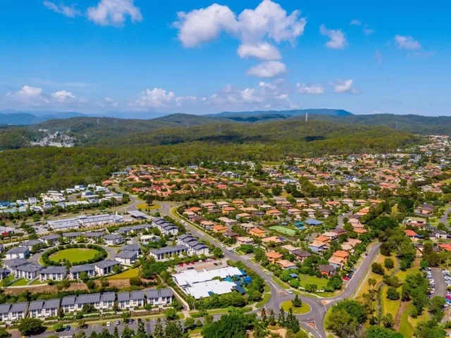 Aerial view of a suburban neighborhood with houses, green lawns, and trees, surrounded by hills and mountains in the distance under a partly cloudy sky.