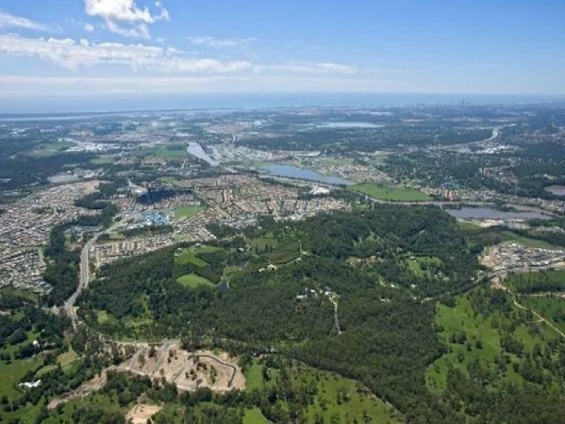 Aerial view of a cityscape with green parks, water bodies, and urban areas under a partly cloudy sky.