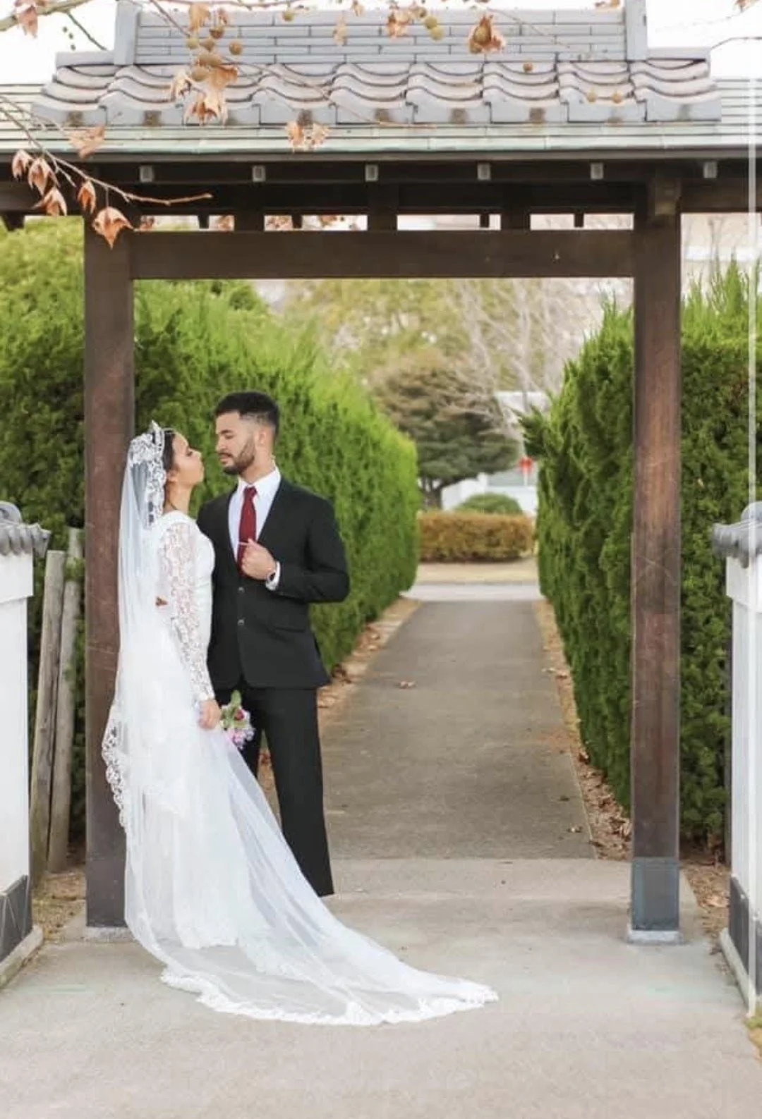 A bride and groom standing under a wooden gate on a pathway, facing each other, with the bride holding a bouquet. The bride is wearing a white lace wedding dress with a veil, and the groom is dressed in a black suit with a red tie. There are green bu