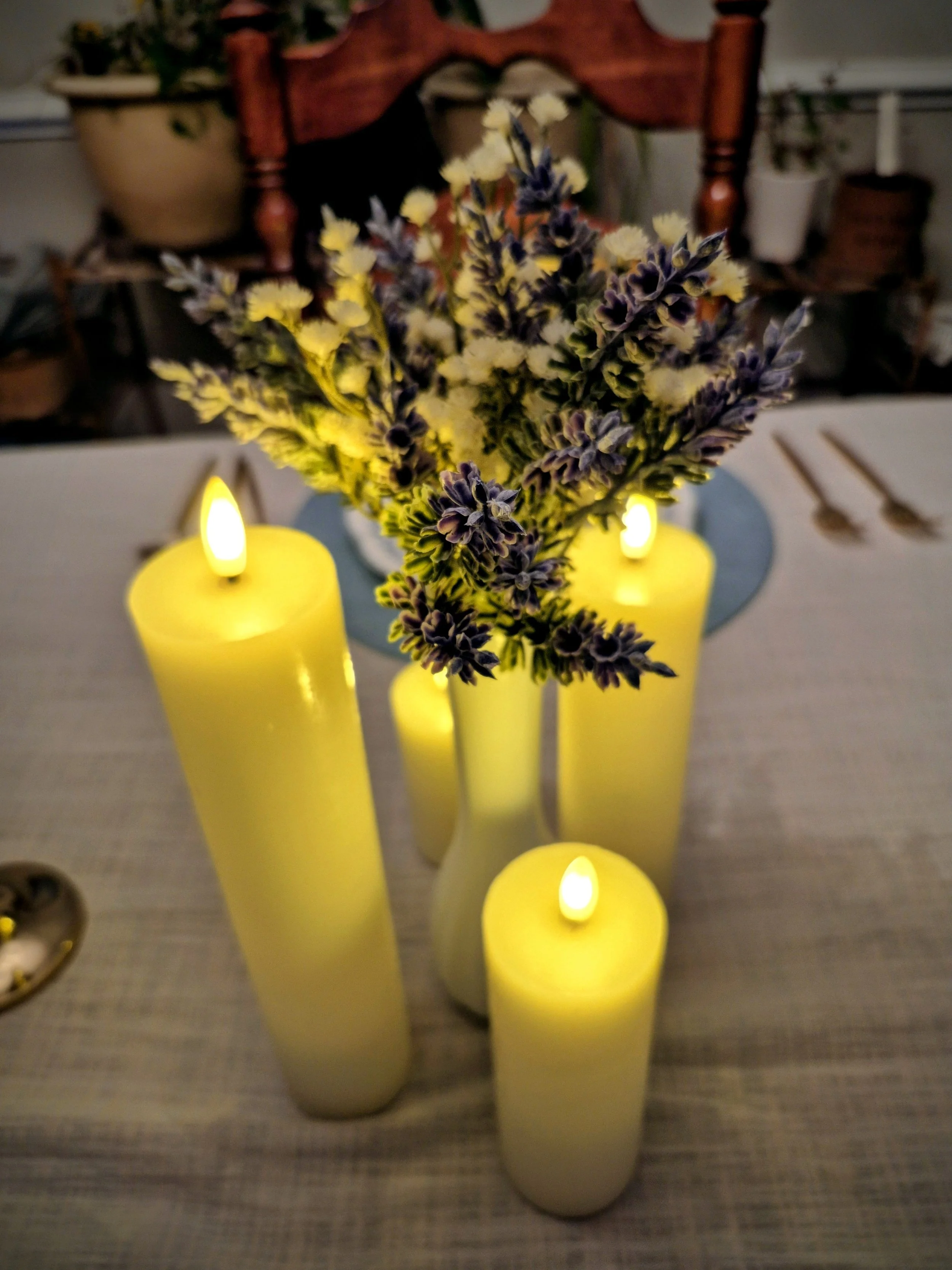 A bouquet of purple and white flowers in a white vase, surrounded by four yellow candles with flickering flames on a table.