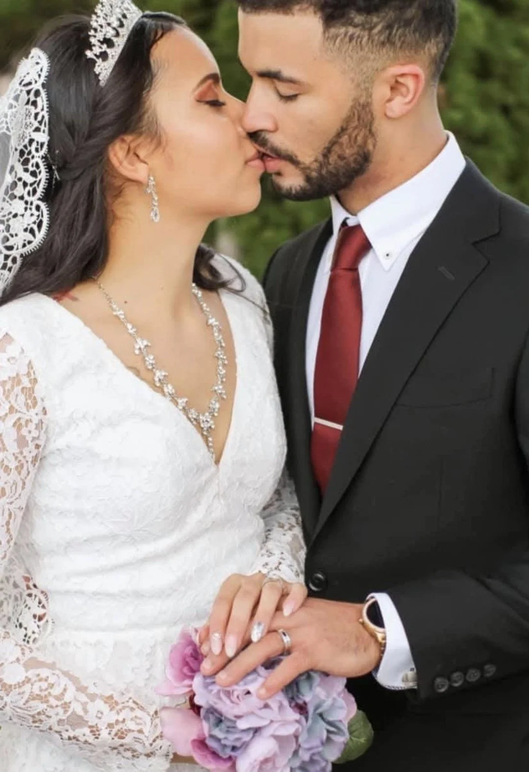 A bride and groom close together, about to kiss, at their wedding. The bride wears a lace wedding dress, a tiara, earrings, a necklace, and holds a bouquet of pink and purple flowers. The groom wears a black suit, white shirt, red tie, and a watch.