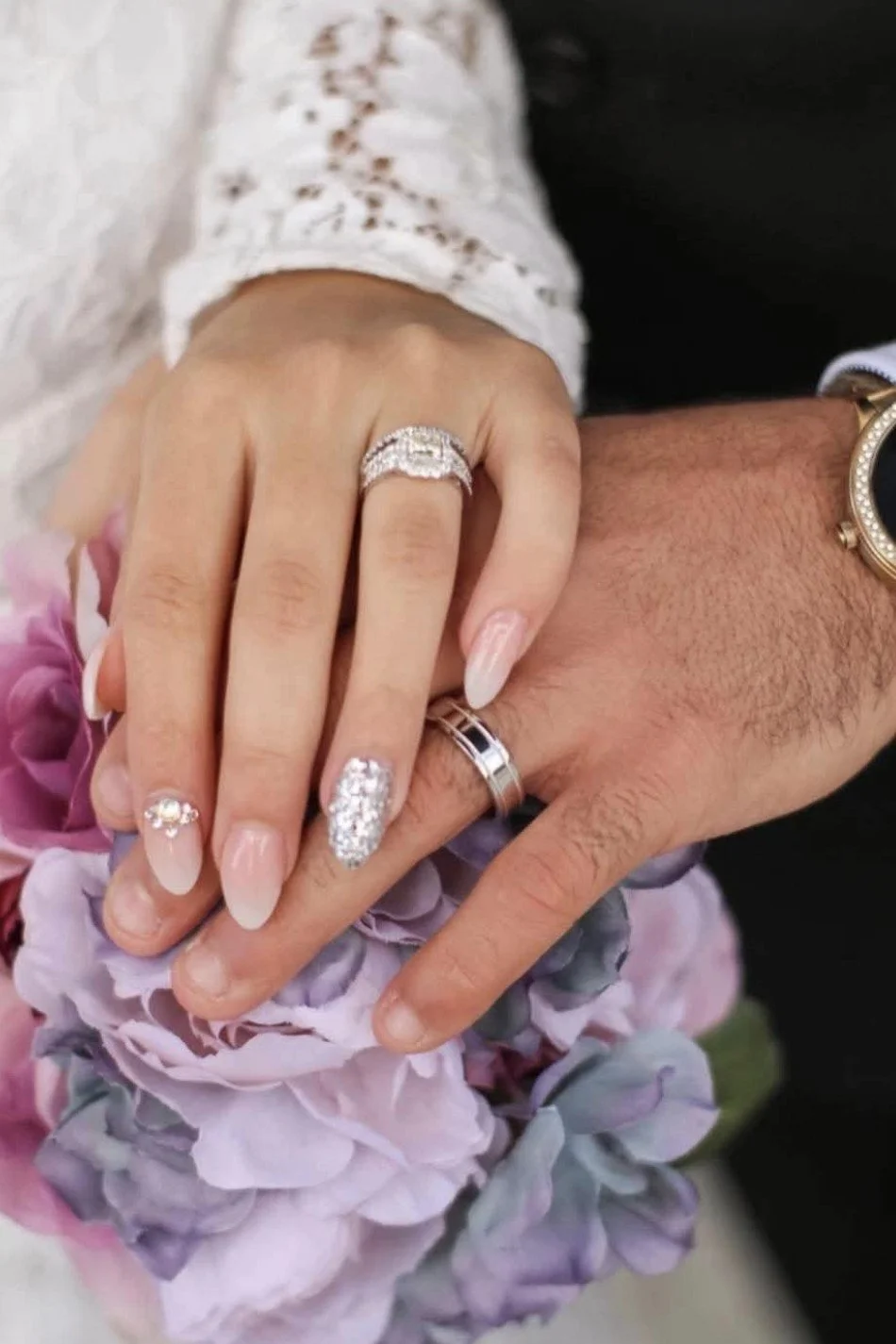 Close-up of a woman's and man's hands with wedding rings, resting on a bouquet of pink and purple flowers.