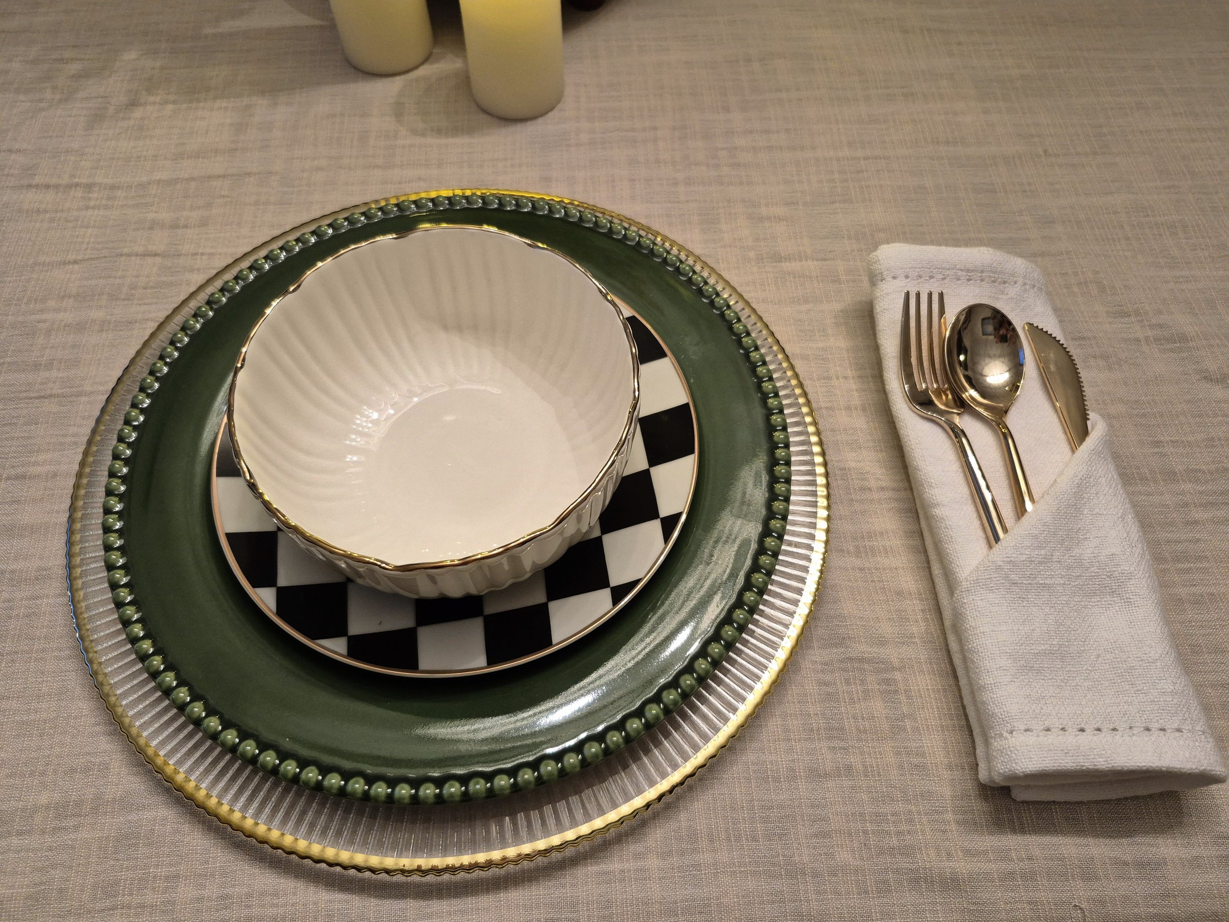 Stacked green, black and checkered plates with a white bowl on top, silverware wrapped in a white napkin on a beige tablecloth.