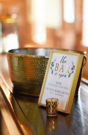 A sign on a bar counter indicating the bar is open, with a small shot glass in front of it. The sign mentions beer, wine, liquor, and frozen margaritas.