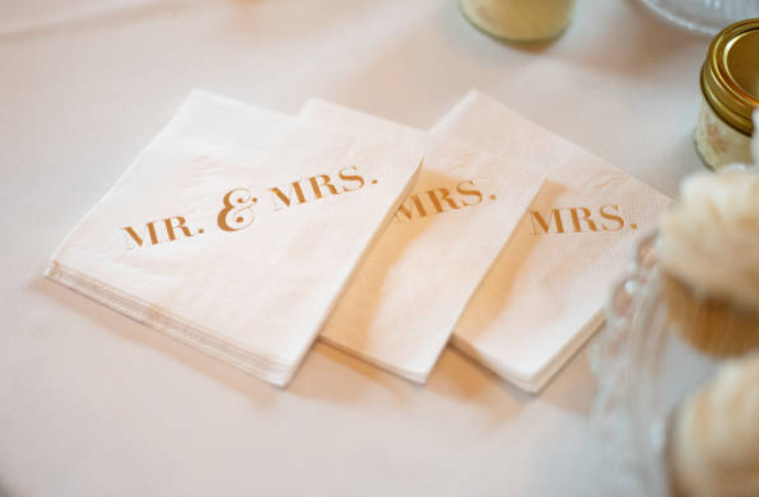 Three white napkins with gold text that say 'Mr. & Mrs.' on a table, alongside jars and blurred decorative items.