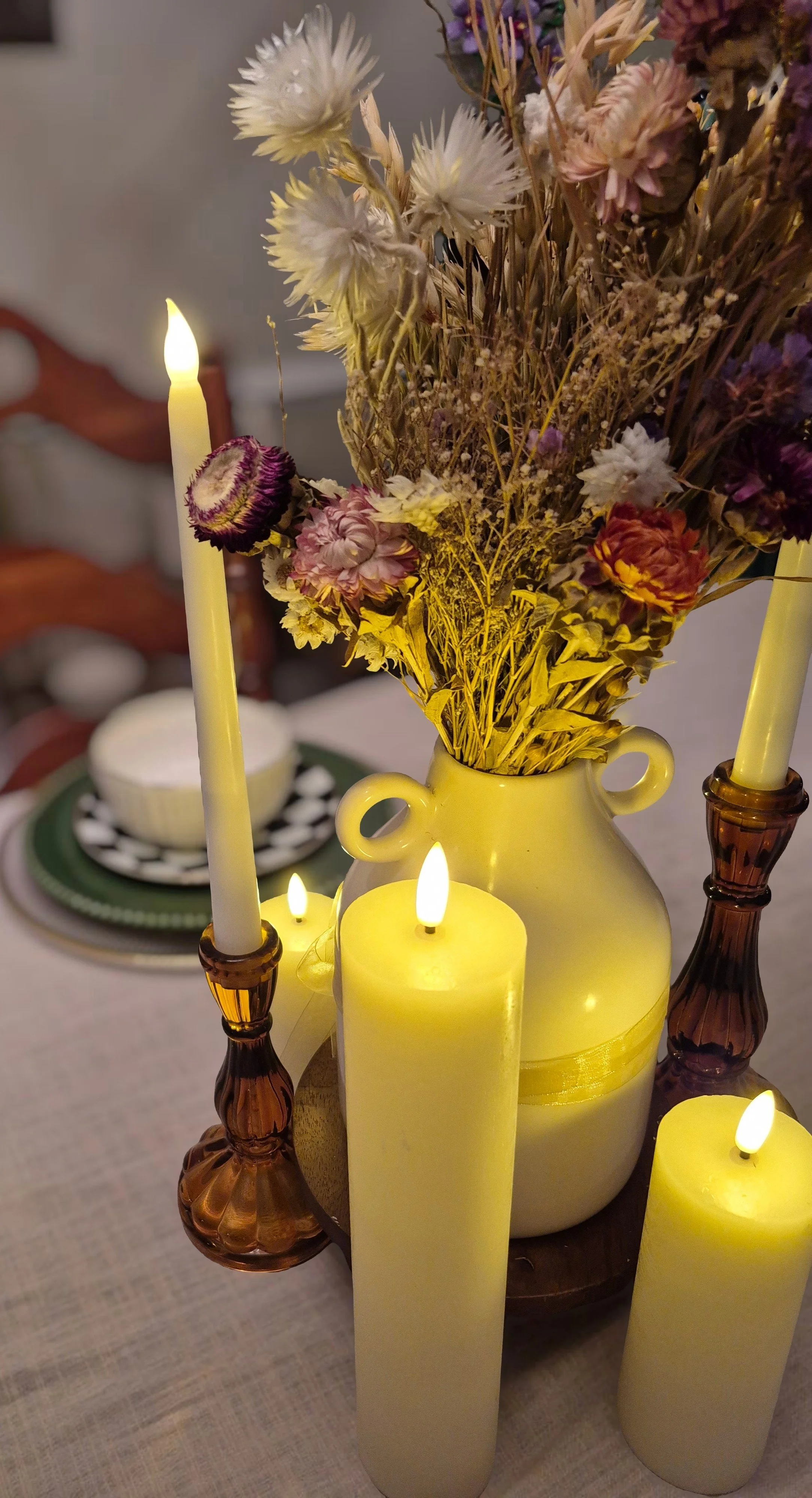 A table centerpiece featuring a bouquet of dried flowers in a yellow vase, illuminated by yellow candles and surrounded by tall candles. In the background, there is a place setting with a bowl on a plate, with a blurred wooden chair visible.