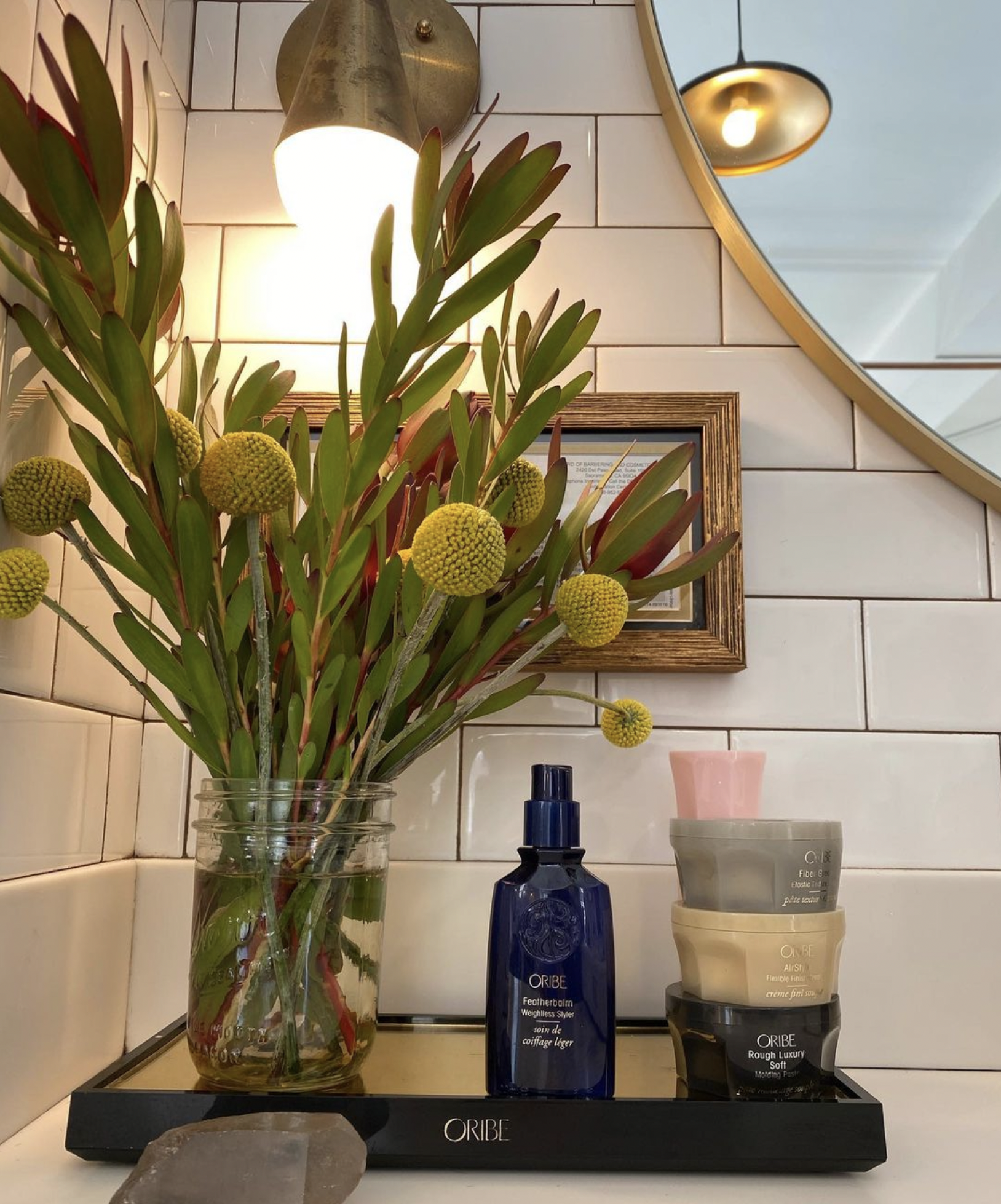 Oribe hair products and fresh florals styled on a salon vanity tray in a modern tile and brass mirror setting.