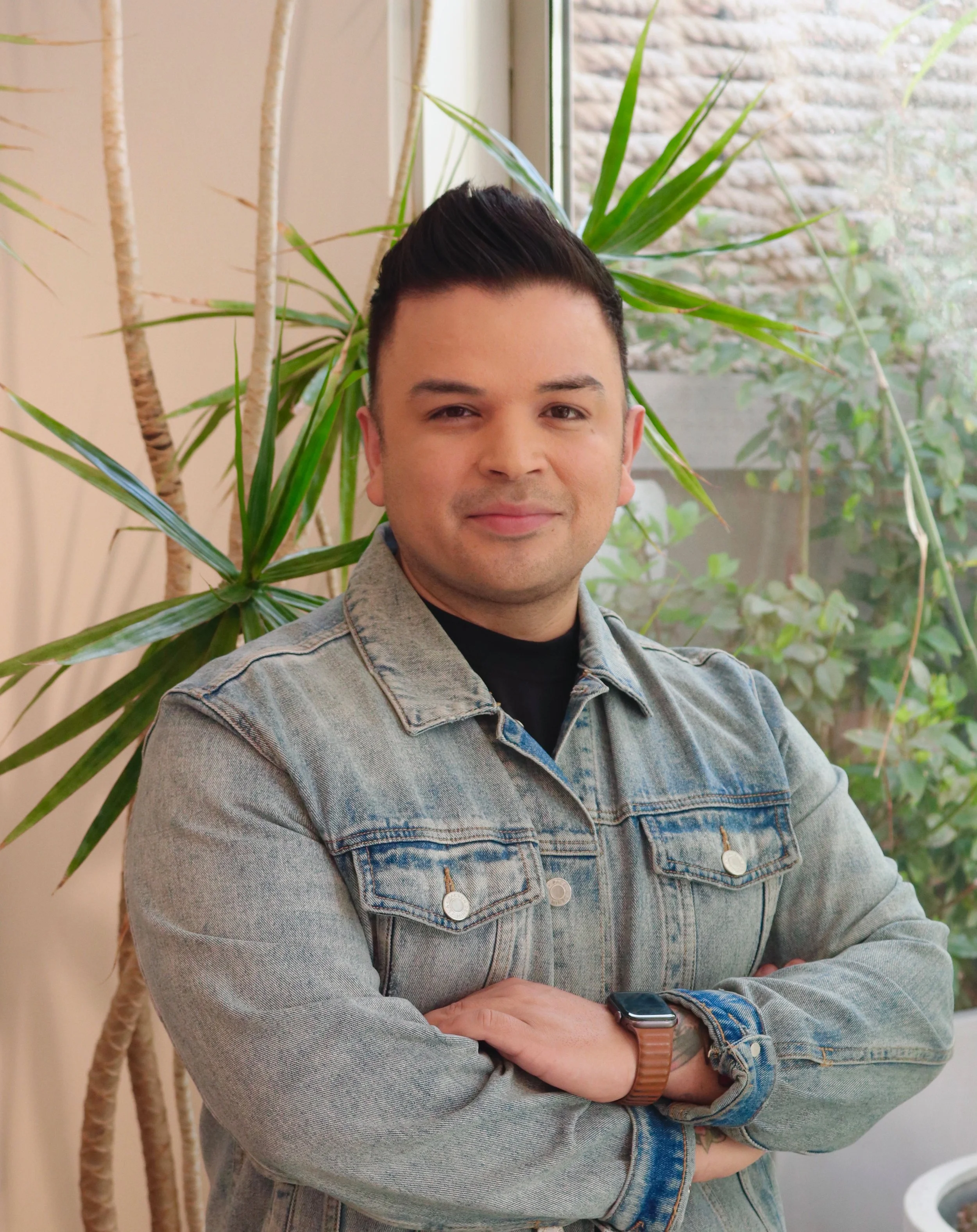 A man with short dark hair, wearing a light denim jacket and black shirt, standing in front of green plants and a window.