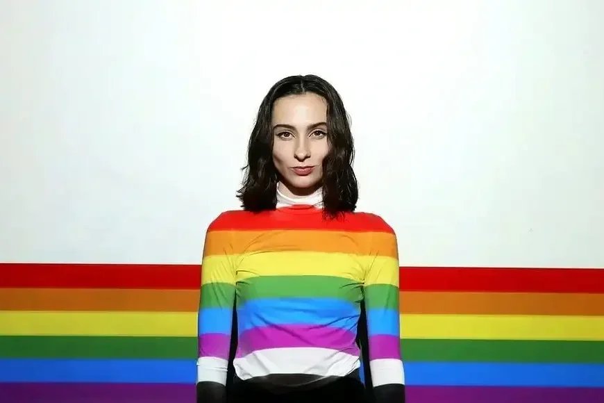 Person wearing a rainbow-striped shirt standing in front of a rainbow backdrop, representing gender-affirming haircut services.