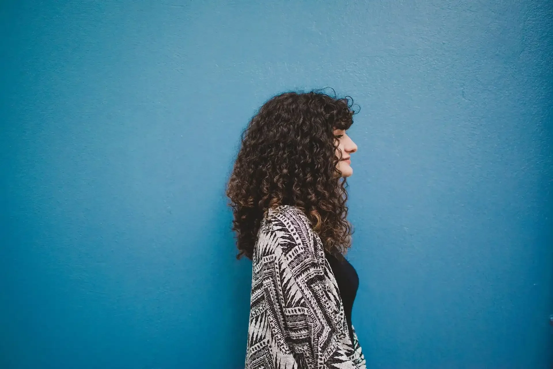 Person with shoulder-length curly hair standing in profile against a blue wall, showcasing natural curl texture.