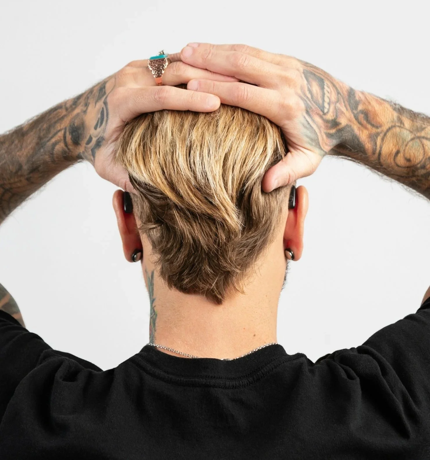 Person with tattoos and earrings, wearing a black shirt, holding their head with both hands, showing short, blonde and brown hair, and wireless earbuds.