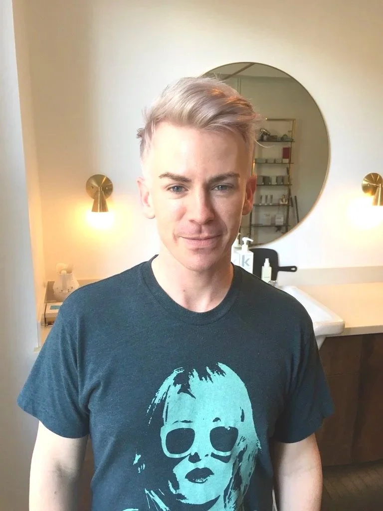 Young person with short blond hair standing in a modern bathroom with round mirror, wall-mounted lights, and shelves with toiletries.
