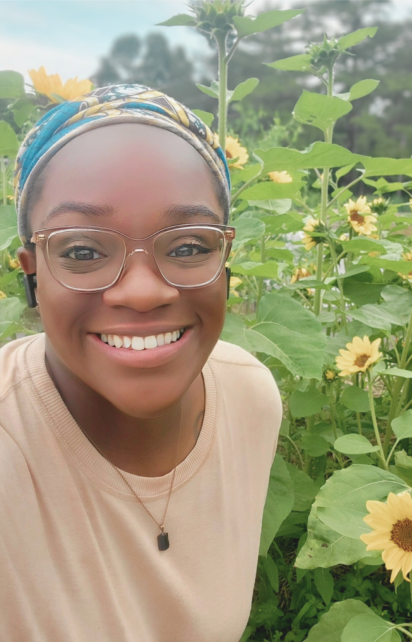Smiling woman taking a selfie in a sunflower field, wearing glasses and a colorful headscarf.