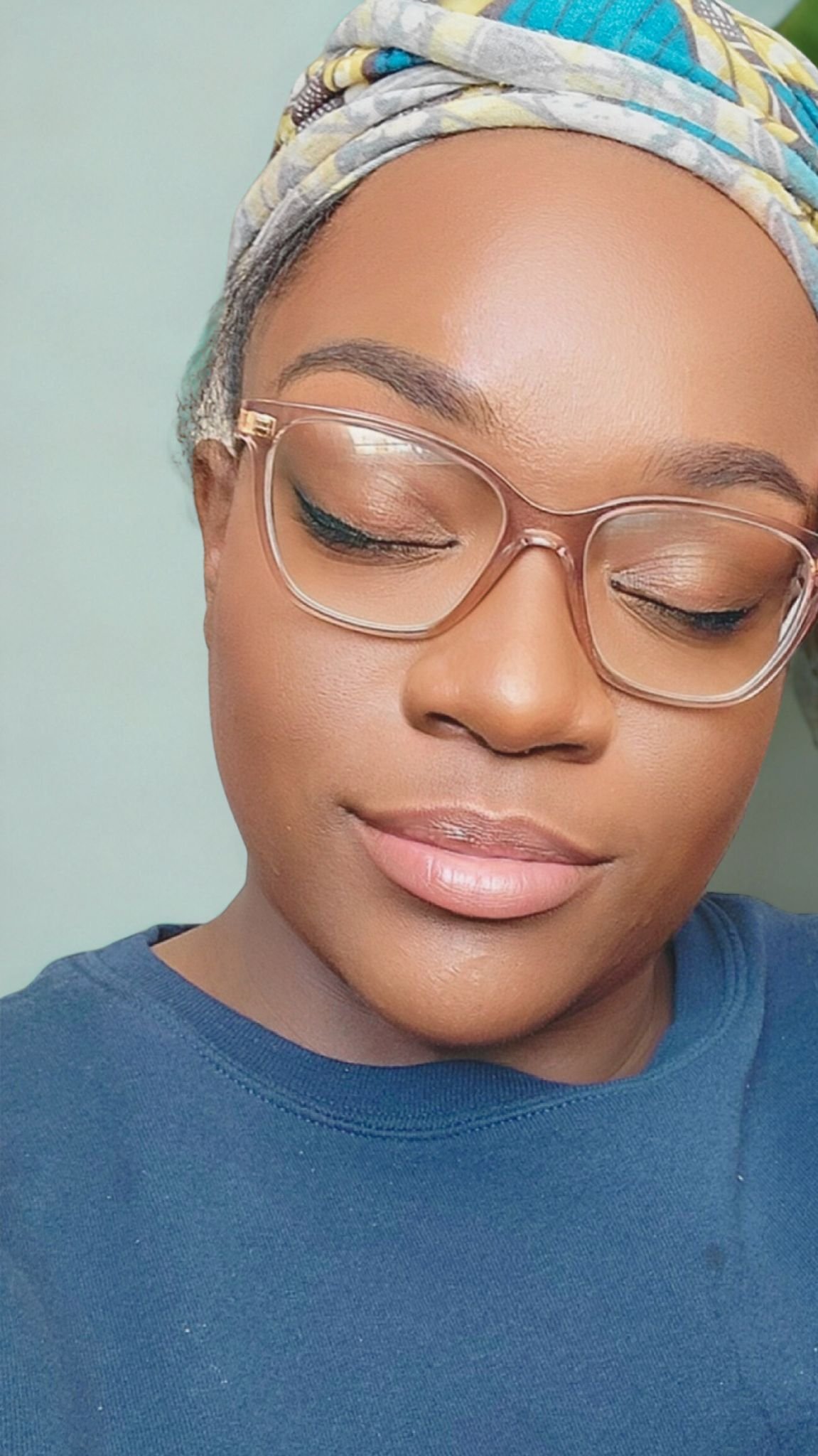 Close-up of a woman with glasses, wearing a colorful headscarf and a blue shirt, with her eyes closed and a peaceful expression.