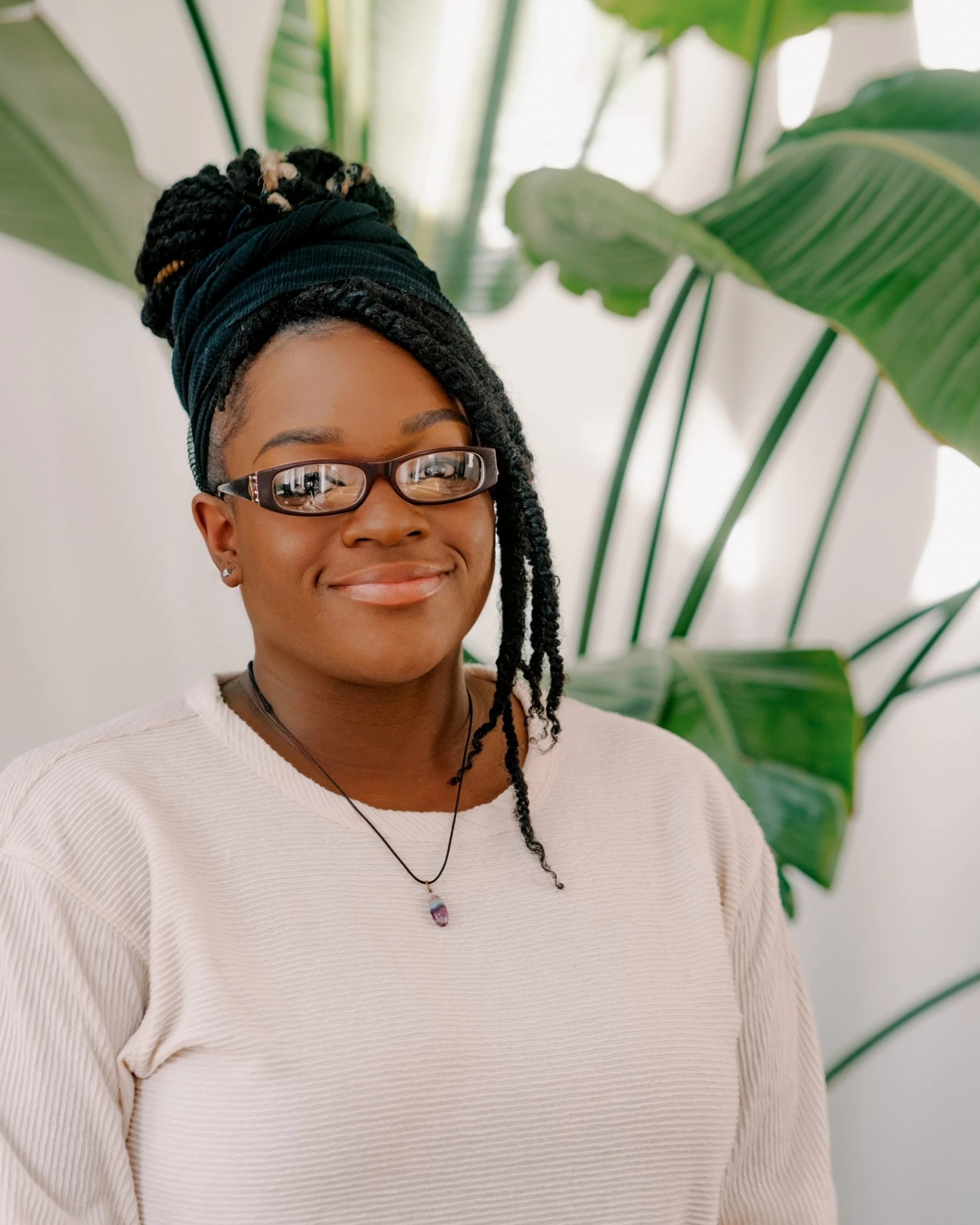 A woman with glasses, braided hair in a bun with a black headwrap, wearing a beige top, standing in front of large green tropical leaves.