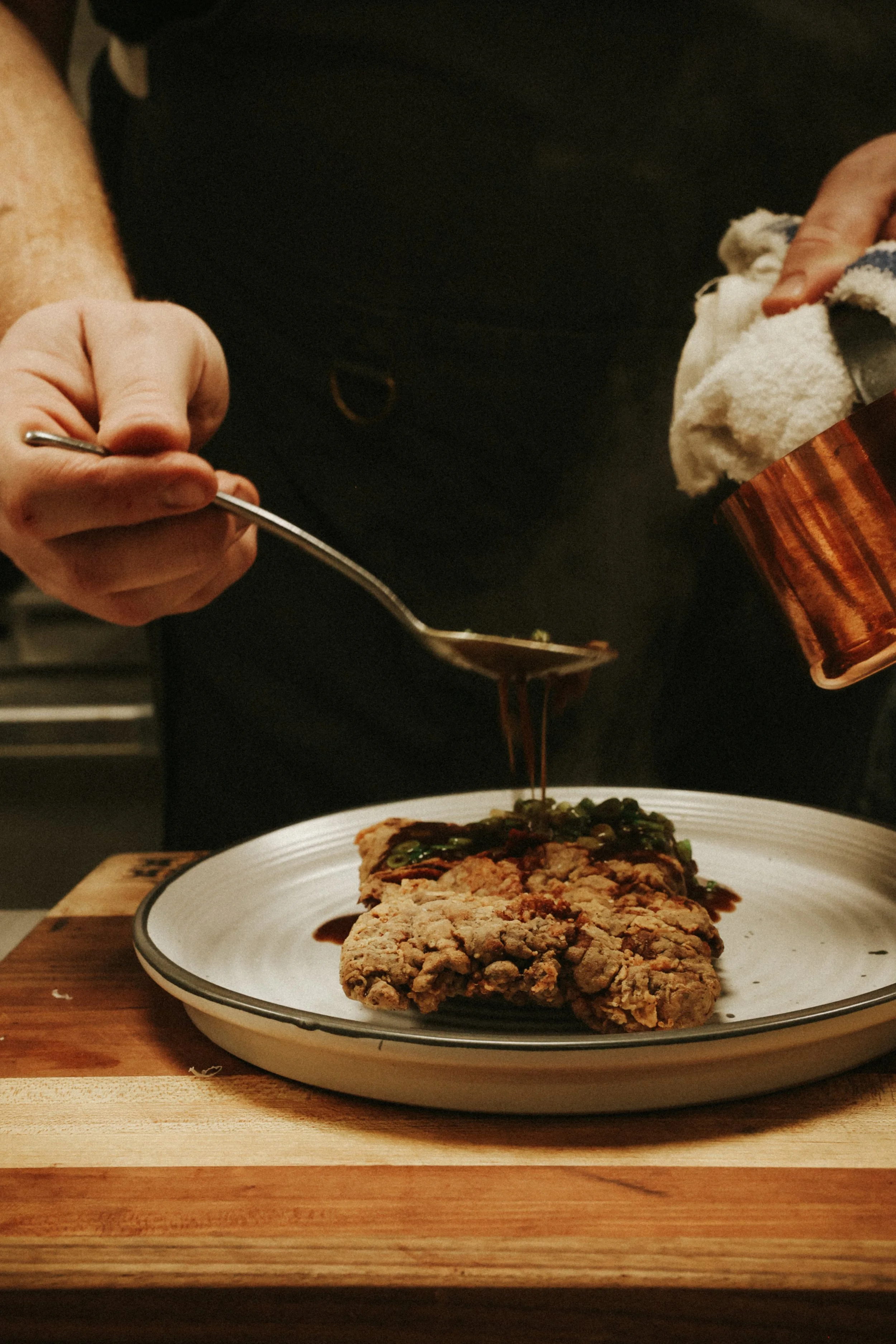 Person pouring gravy over a slice of meatloaf on a plate, holding a spoon in one hand and a towel in the other.