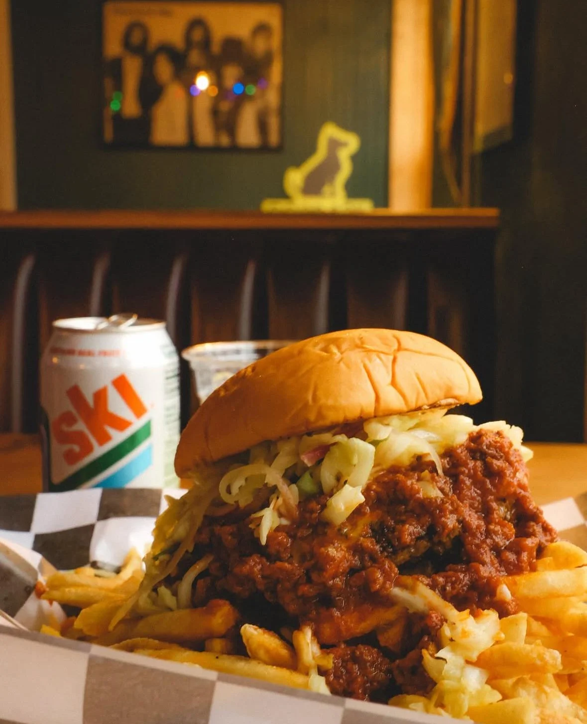 Close-up of a sloppy joe sandwich with shredded lettuce and fries on checkered paper, with a can of SKI soda in the background on a wooden table.