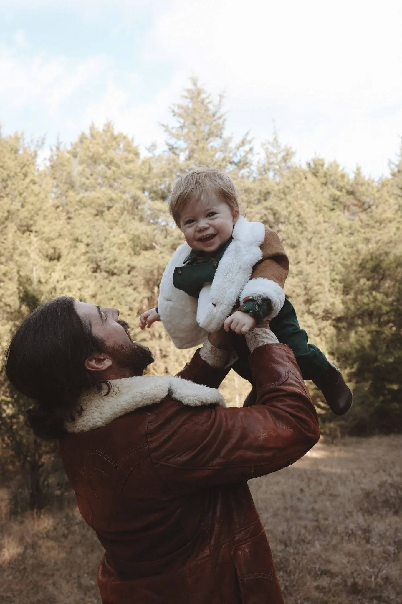 A man with a beard and brown leather jacket lifts a smiling young child with light brown hair into the air outdoors, trees in the background, with a bright sky.