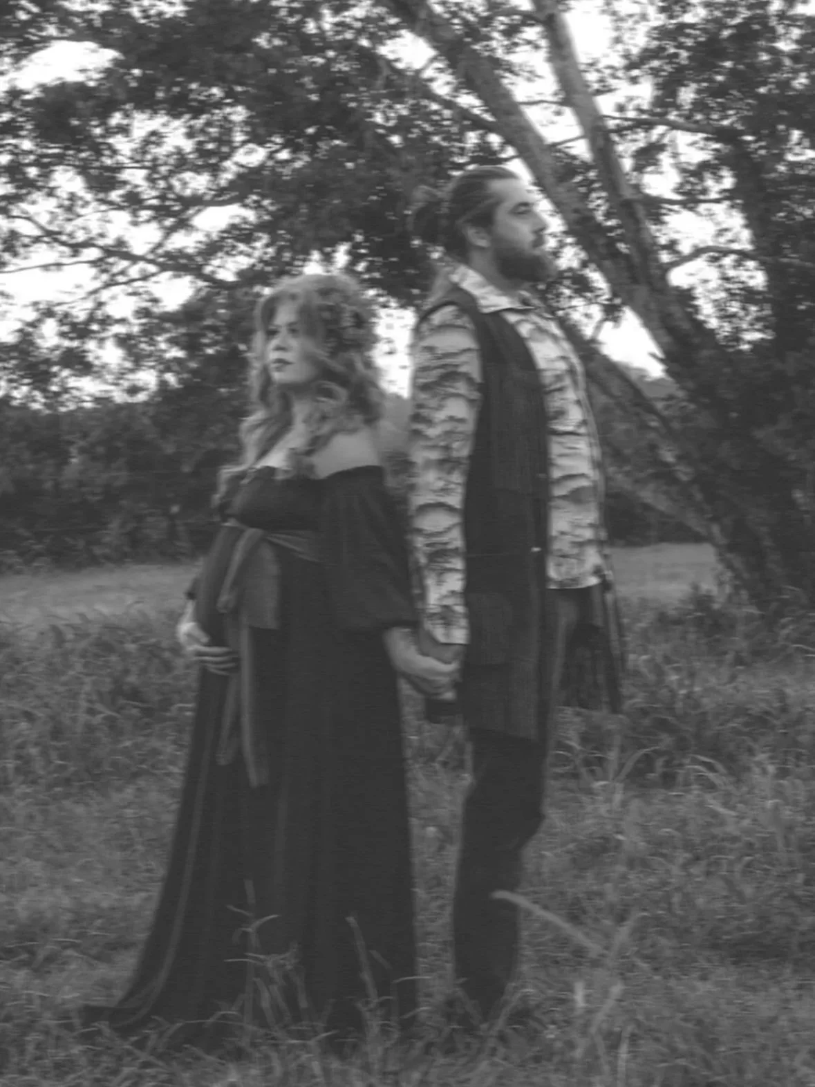 A black and white photo of a man and woman holding hands outdoors near trees, with both facing sideways.