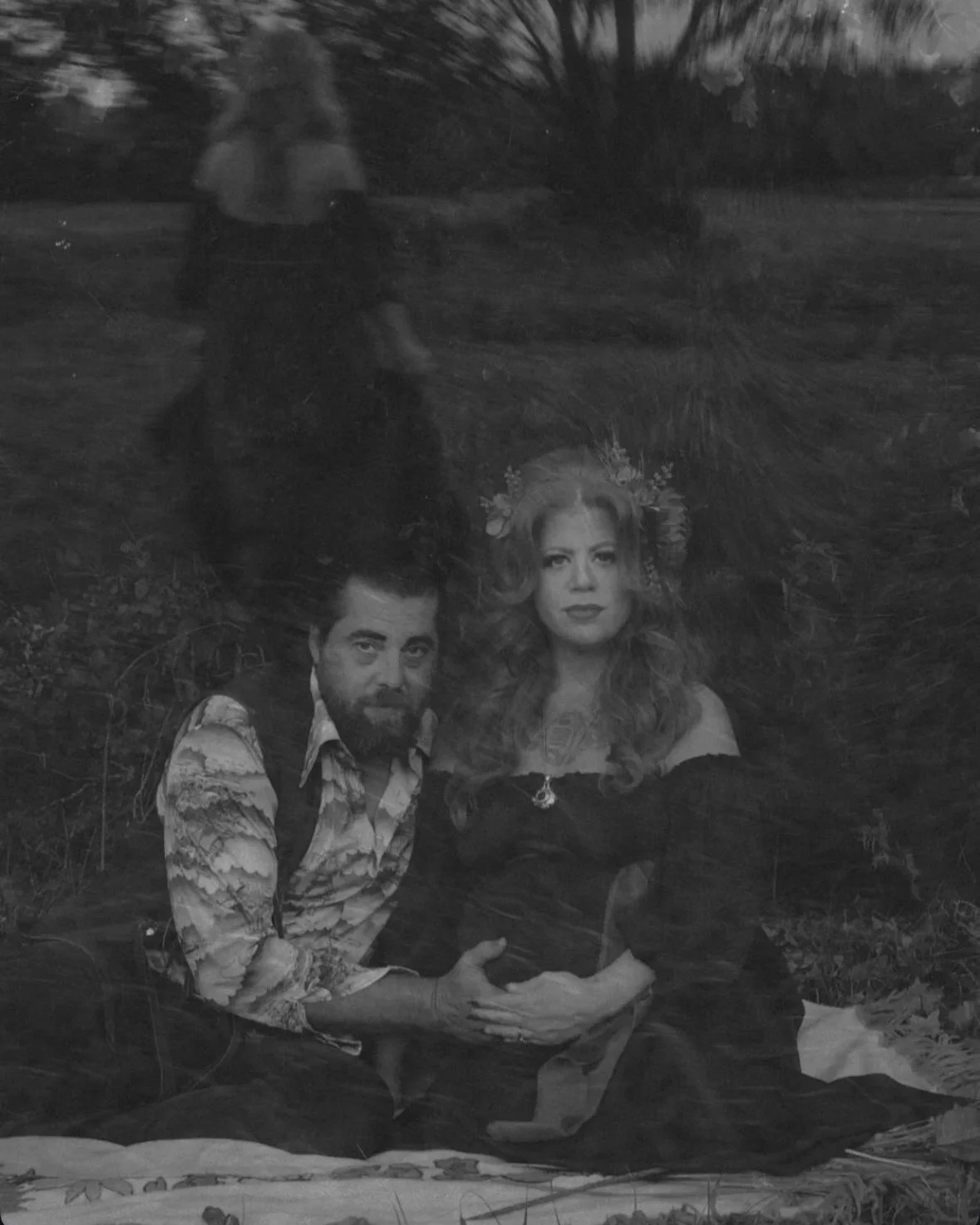 A black and white photo showing a woman and a man sitting on the ground with a tree and stars in the background. The woman is wearing a floral crown and the man has a beard and is wearing a patterned shirt.