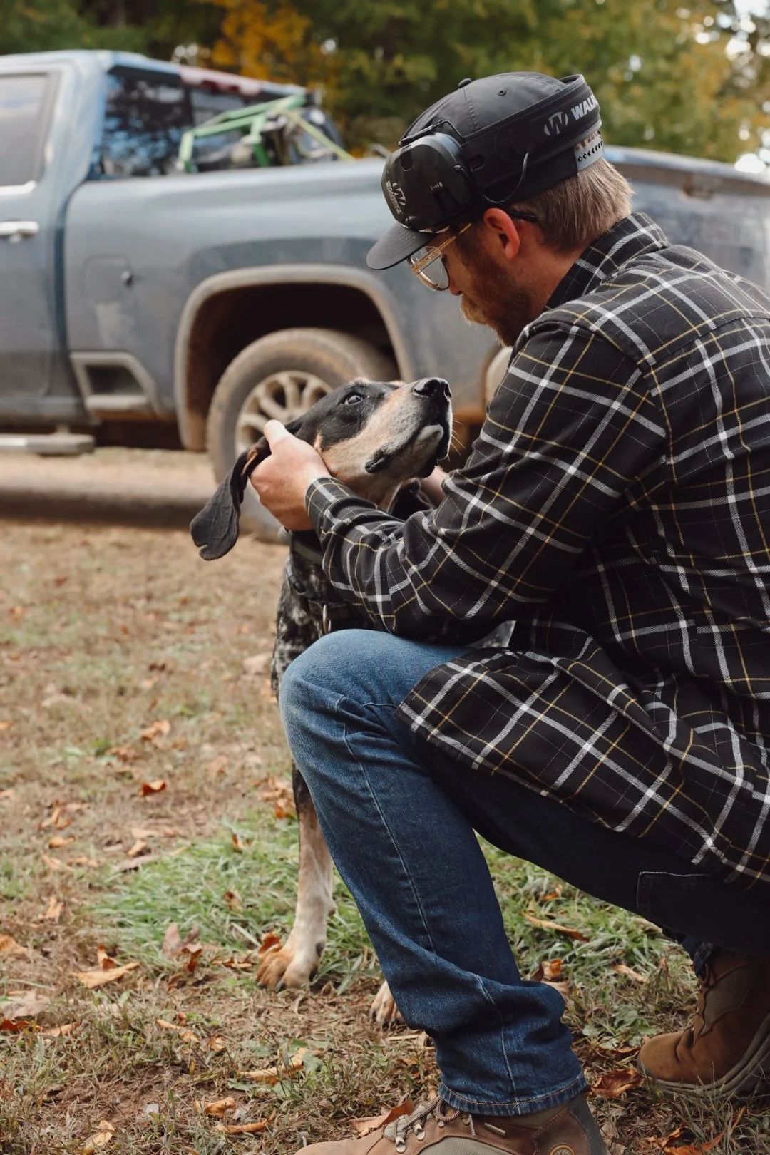 A man wearing a plaid shirt, jeans, and a helmet kneels and gently holds a large black and white dog by the face, with a truck and autumn trees in the background.