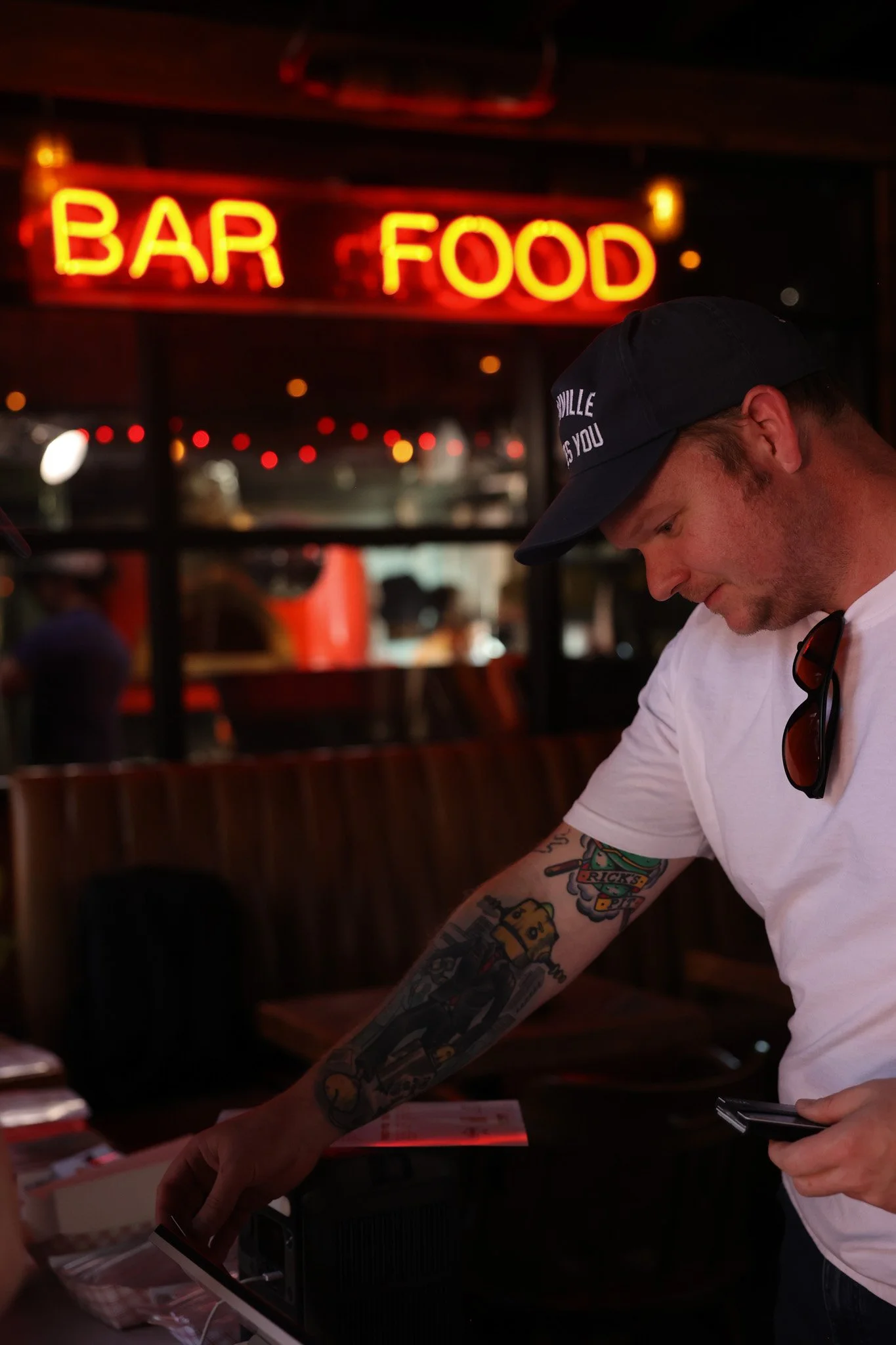 A man wearing a dark baseball cap, white t-shirt, and sunglasses hanging from his collar, with tattoos on his arm, stands inside a bar or restaurant with a glowing red neon sign that reads 'BAR FOOD' behind him.