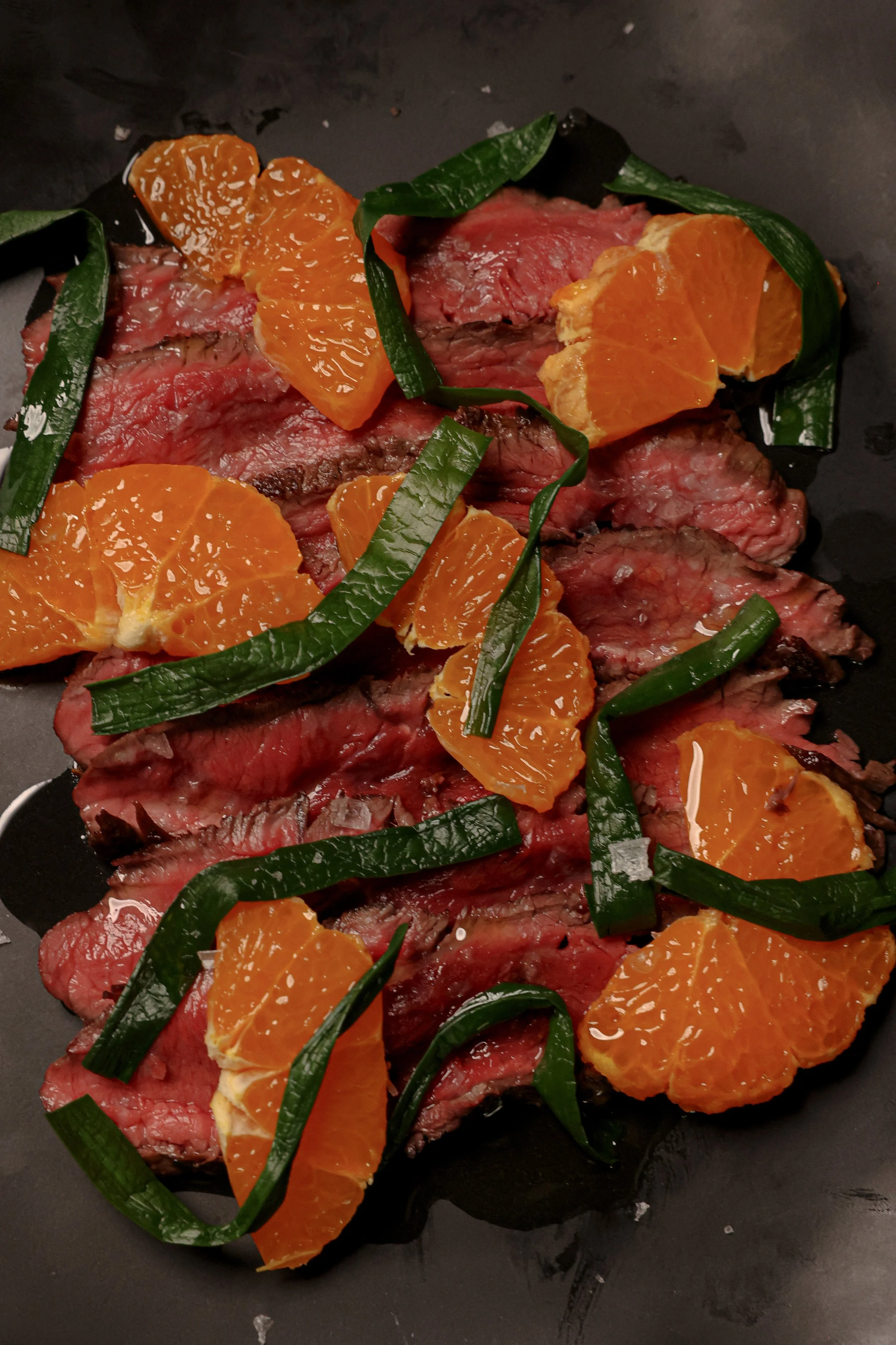 Sliced cooked beef steak topped with orange segments and green sliced herbs.