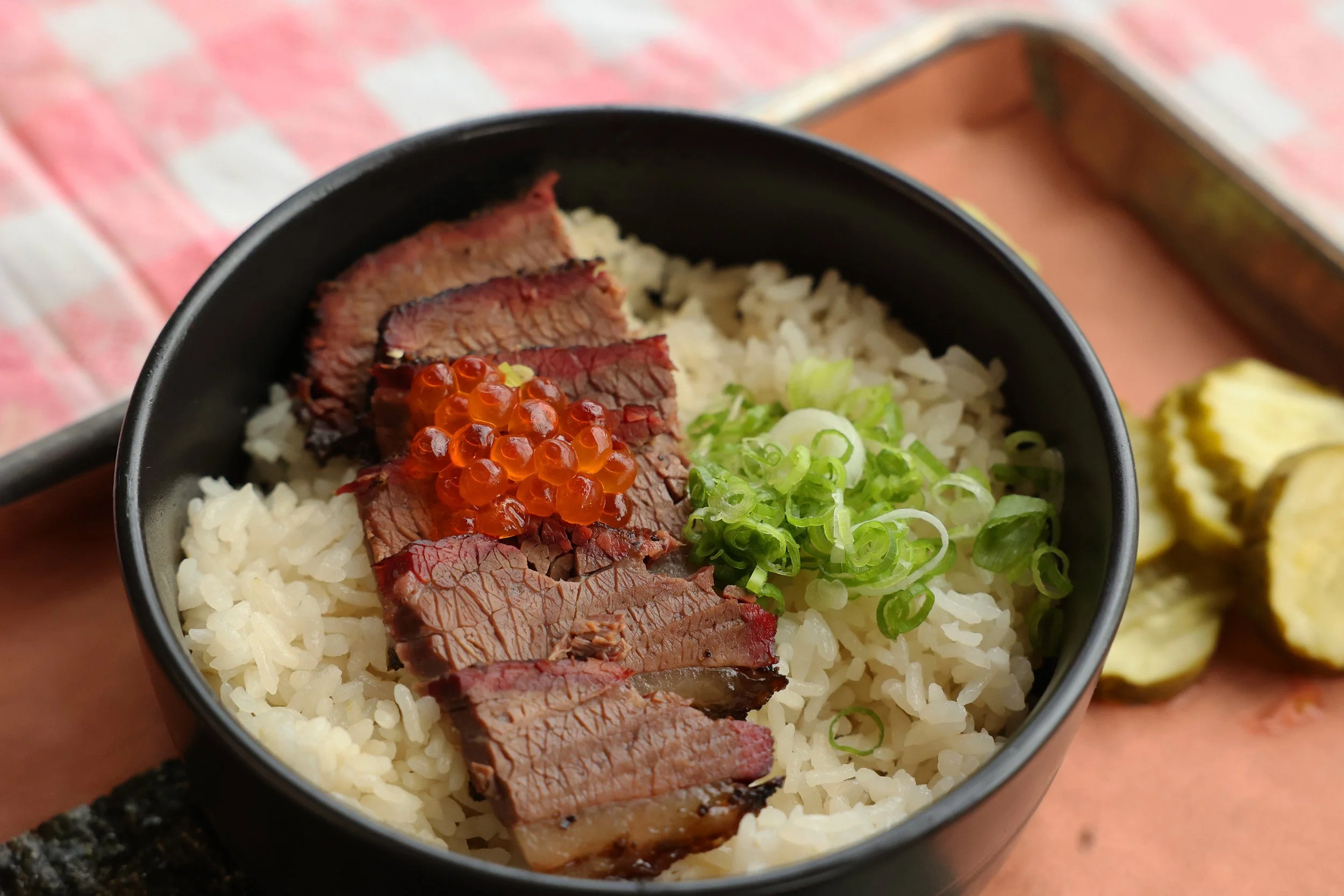 A bowl of sushi rice topped with slices of beef, orange fish roe, chopped green onions, and accompanied by sliced pickles on the side.