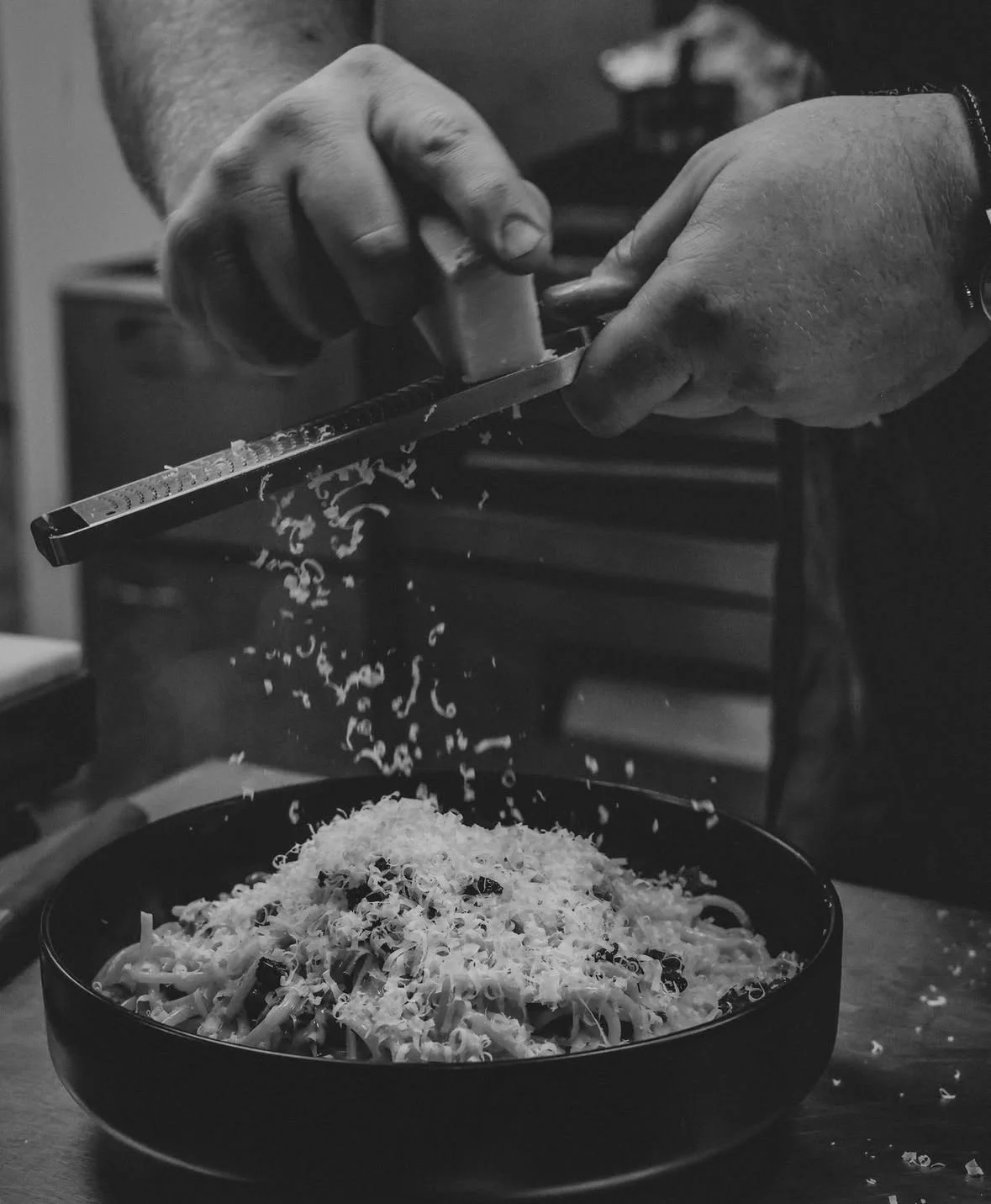 A person grating cheese over a bowl of pasta in a black and white photo.