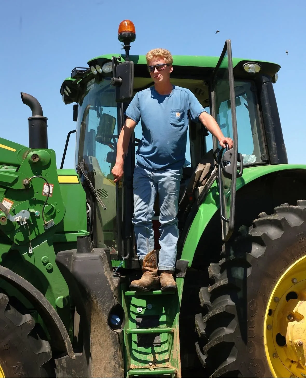 A man standing on the steps of a green tractor, wearing sunglasses, a blue t-shirt, jeans, and boots, with a clear blue sky in the background.