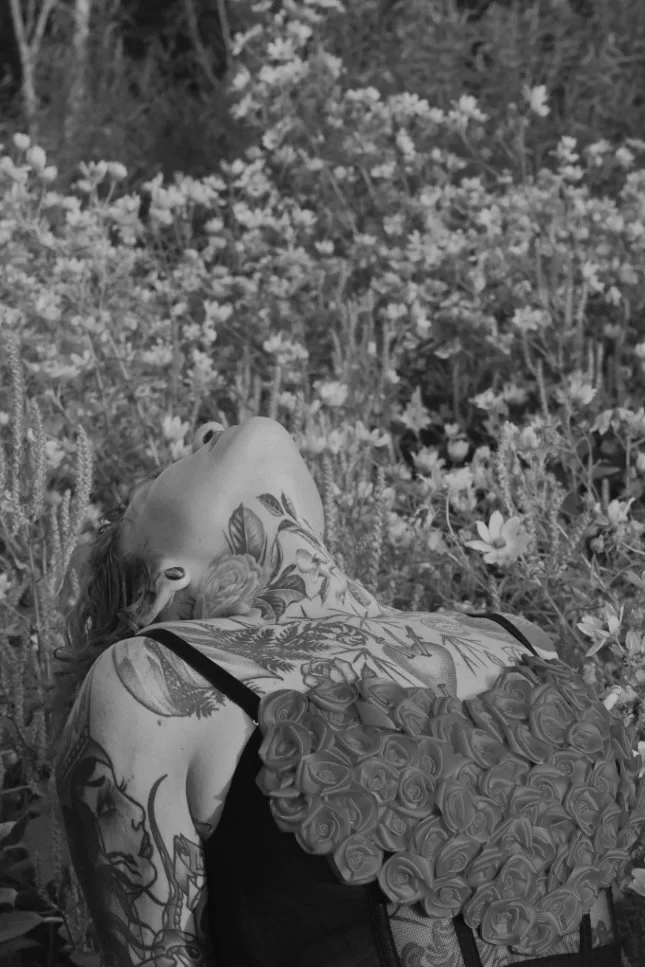 A woman with tattoos on her neck and arms lying in a field of flowers, wearing a dress with a rose pattern.