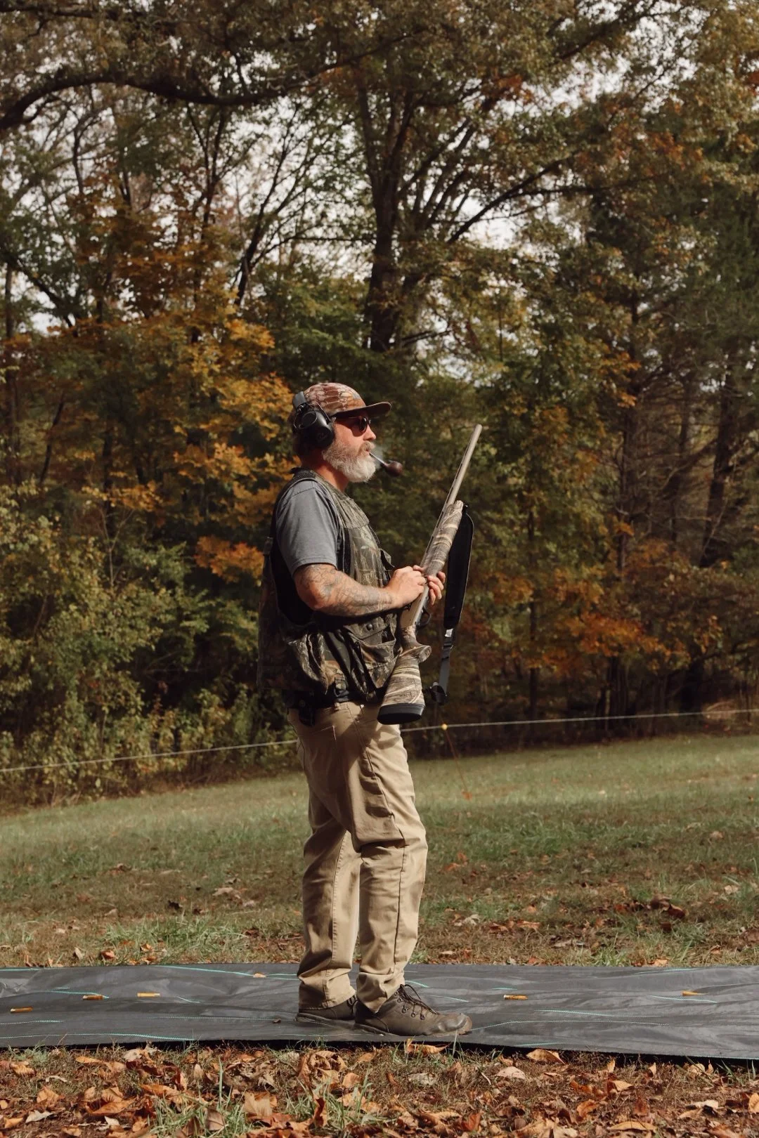 A man with gray hair and beard, wearing a camouflage cap, sunglasses, headphones with microphone, and outdoor clothing, standing on a mat in a wooded area during autumn, holding a rifle, possibly engaging in shooting or hunting activities.
