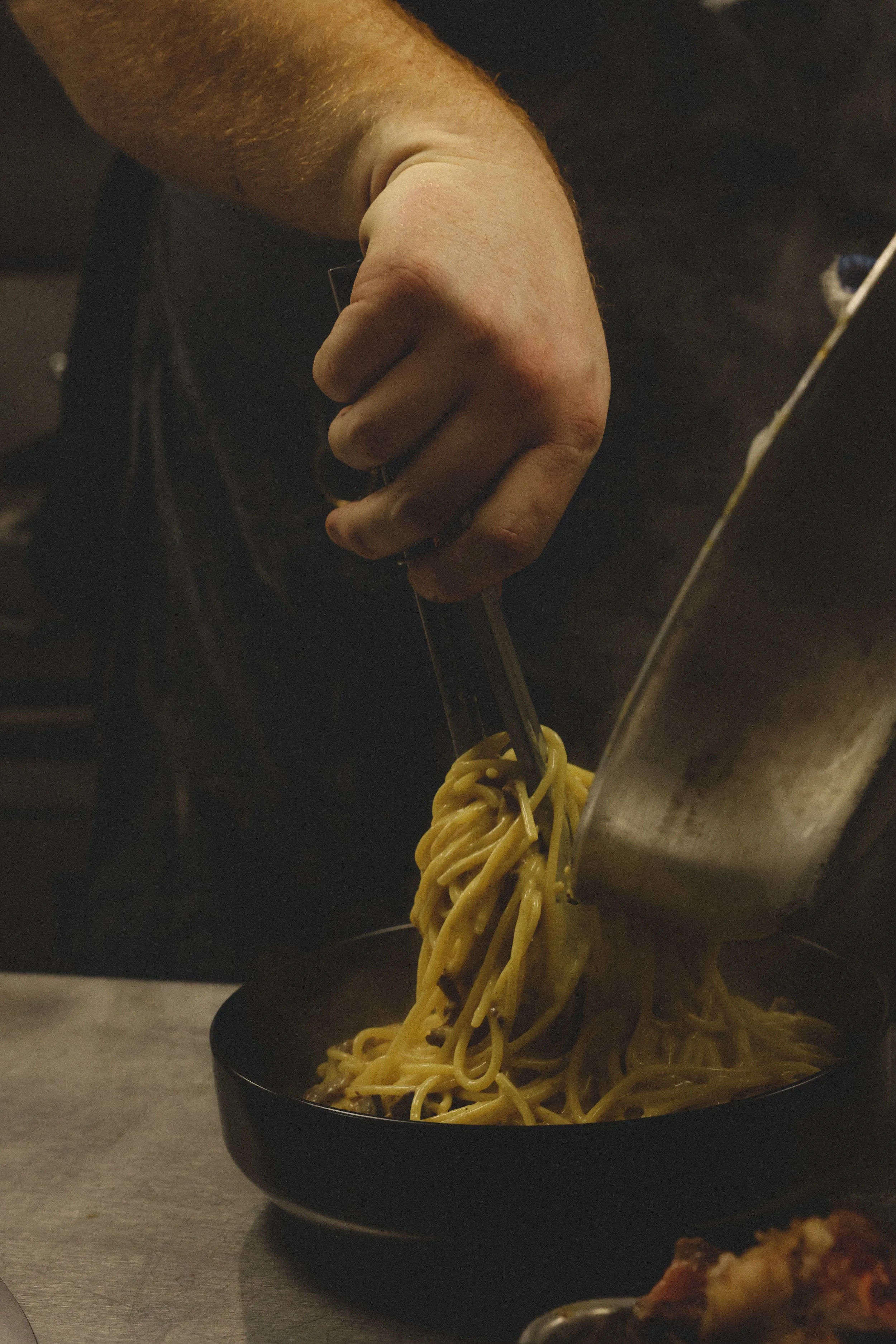 Person using tongs to serve spaghetti noodles into a black bowl.