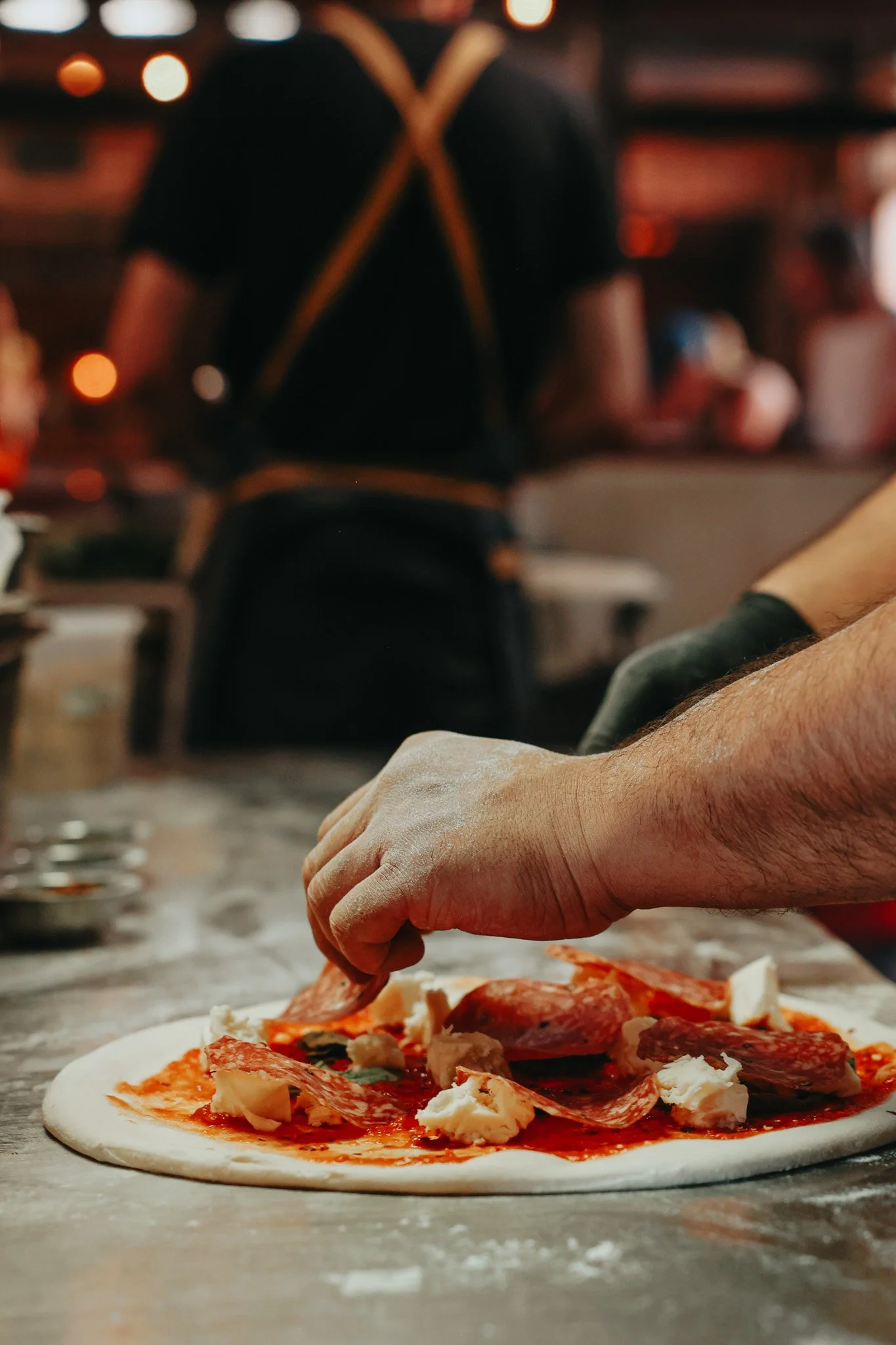 A person adding toppings to a pizza dough with pepperoni, cheese, and tomato sauce in a pizzeria.