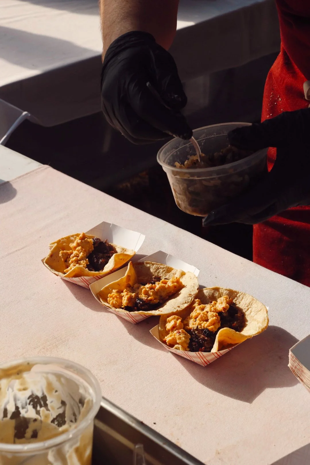 Three mini tacos filled with meat and cheese on a pink table, with a person preparing more fillings in a plastic container and a cup of ice cream in the foreground.