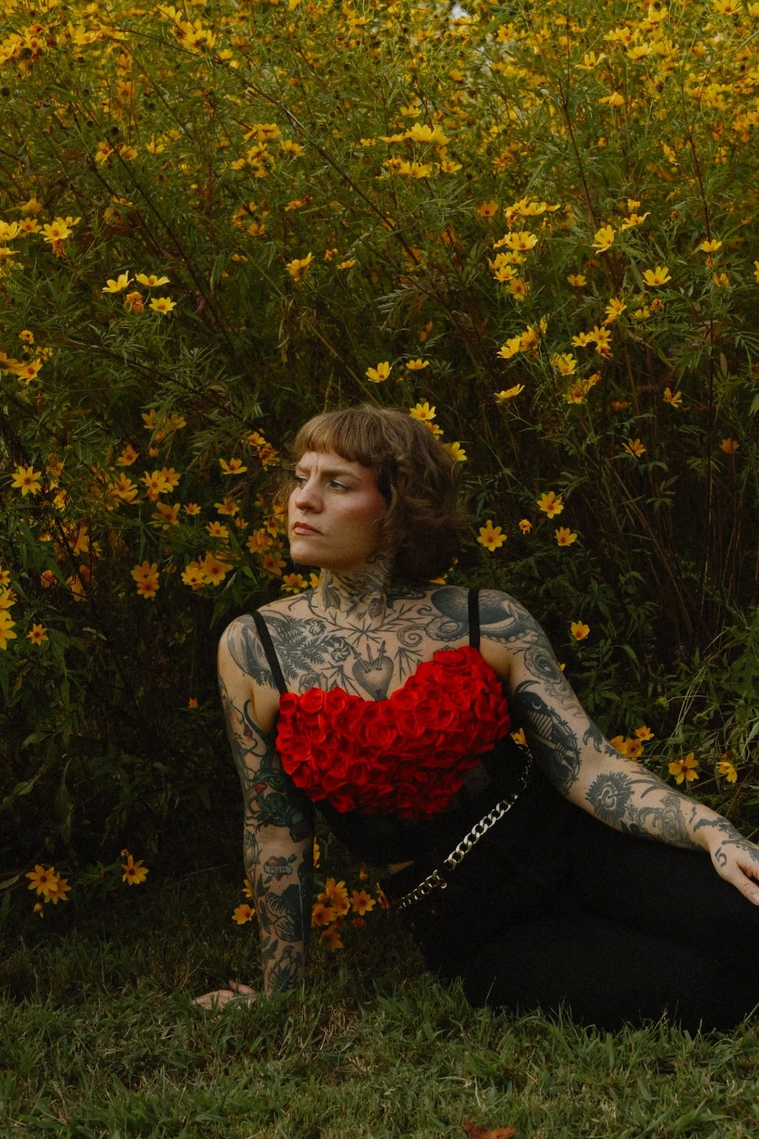 A woman with tattoos on her arms and chest sits on grass in front of yellow flowers, wearing a black dress with a red heart-shaped top made of roses and a chain belt, looking to the side.