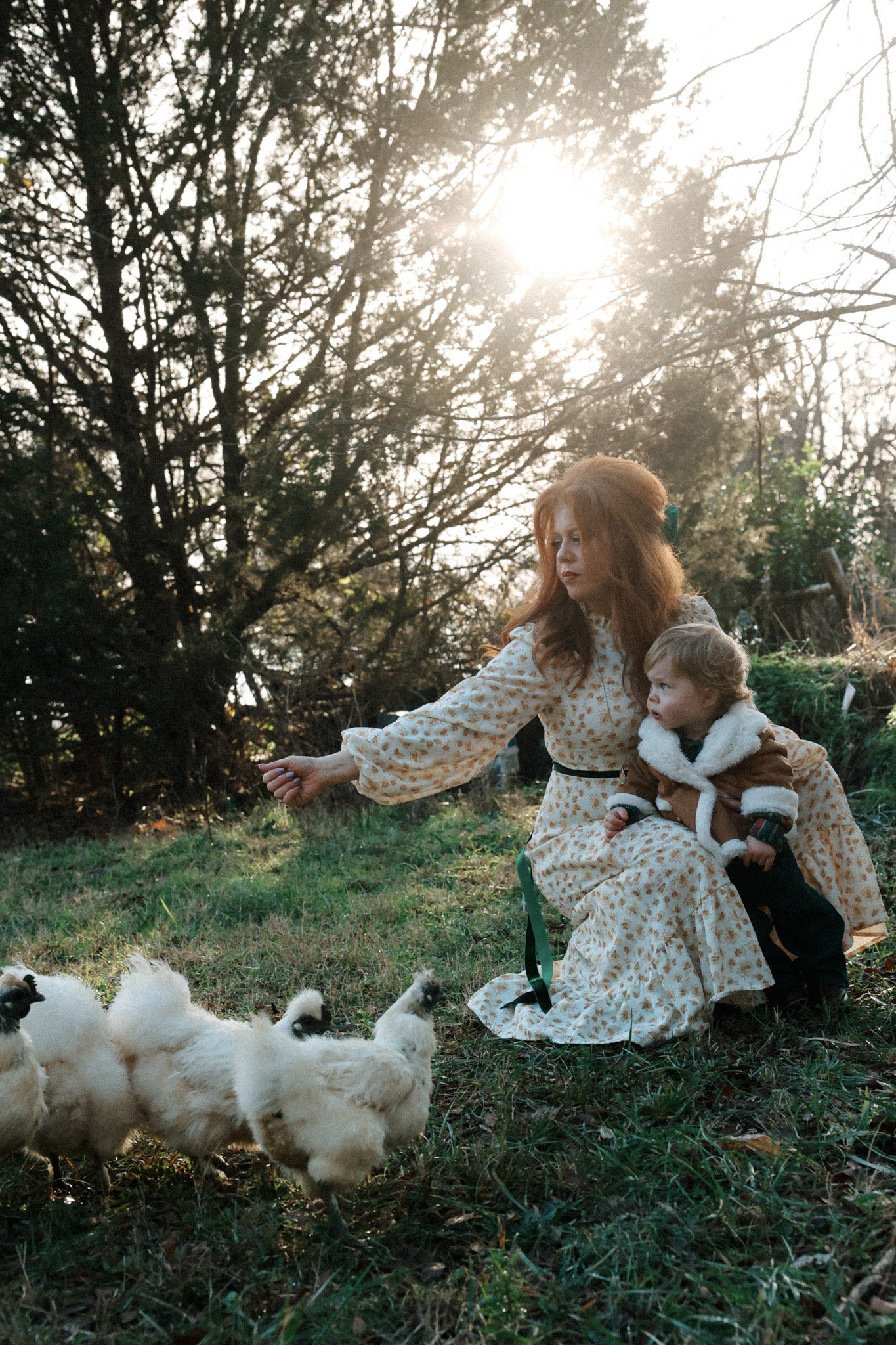 A woman with red hair wearing a long, patterned dress sitting on the grass with a young boy. The woman is reaching out to a group of small, fluffy white chickens. The scene is outdoors with trees and sunlight in the background.