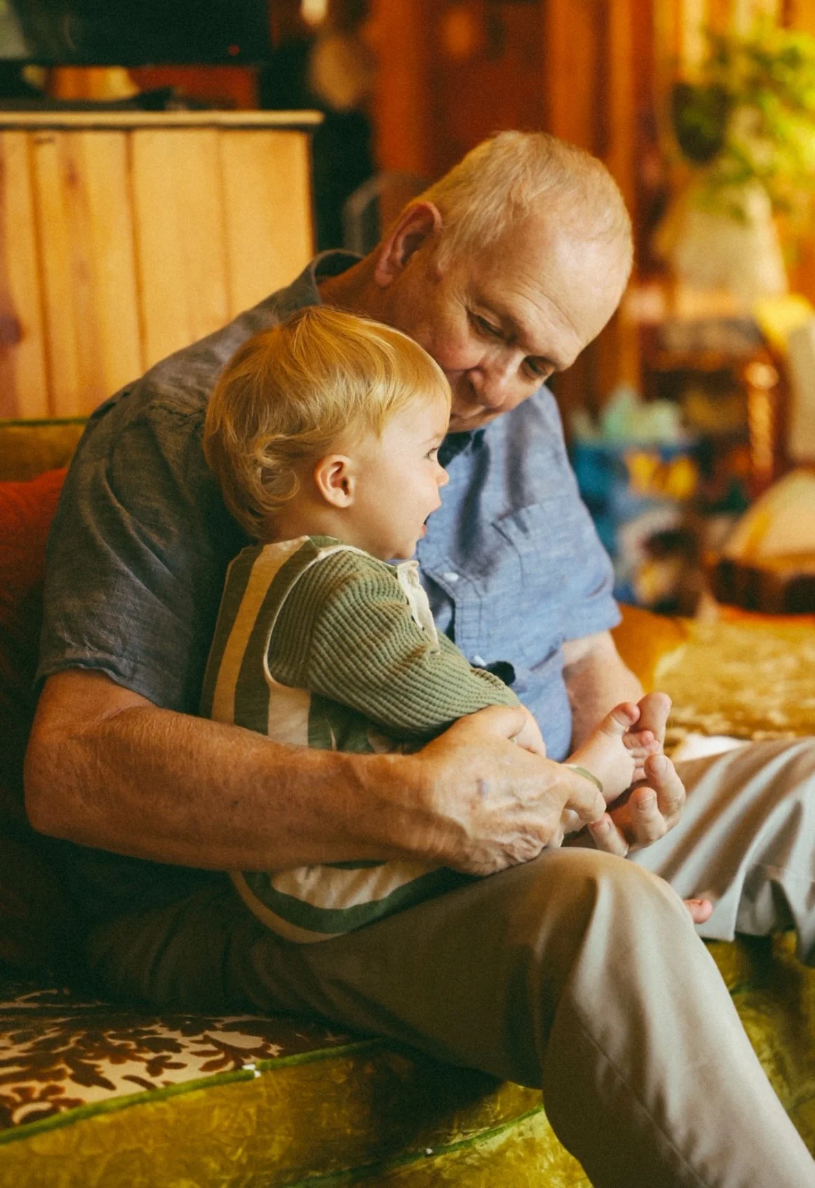 An elderly man with a young child sitting on his lap, playing with their hands together in a cozy, warmly lit room.