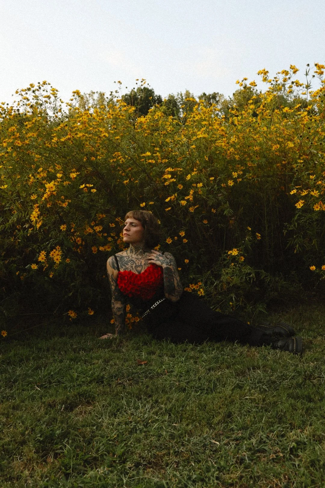 A woman with tattoos, short brown hair, wearing a red top with ruffles, black pants, and boots, sitting on the grass in front of a large yellow-flowered bush, with a neutral expression, looking to the side.